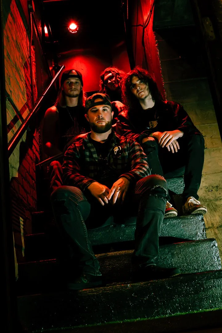 Four young men sitting on dark, wet stairs in a dimly lit environment with red lighting, with brick walls and colorful posters in the background.