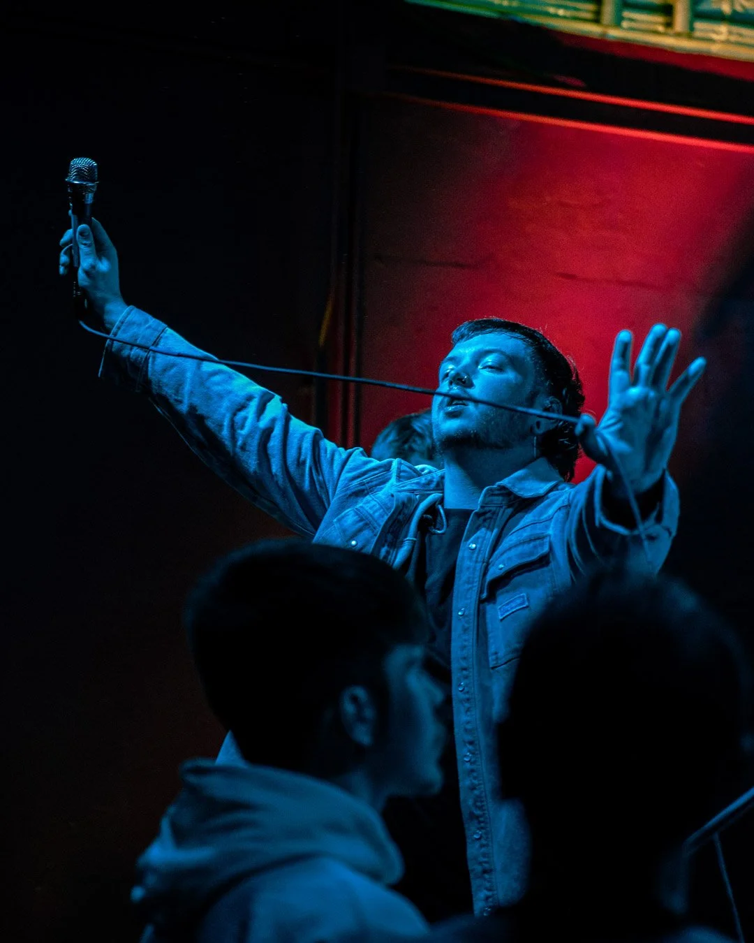 A man holding a microphone stands on stage, with his eyes closed and arms raised, illuminated by blue and red stage lights, with a crowd in front of him.