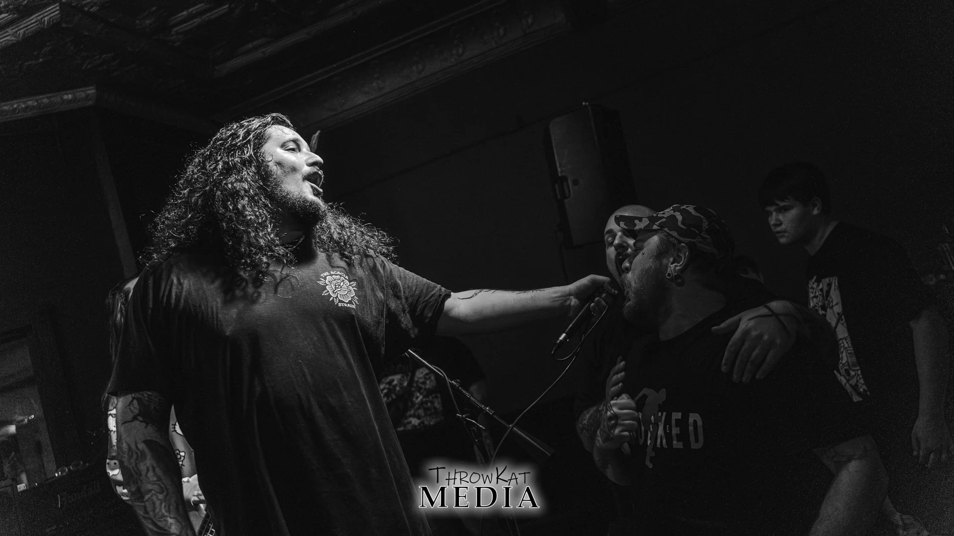 A black and white photo of a live musical performance. A man with long, curly hair and tattoos on his arm is singing into a microphone, with his hand on another man's shoulder. The second man is wearing a camouflage cap and earrings, holding a microp