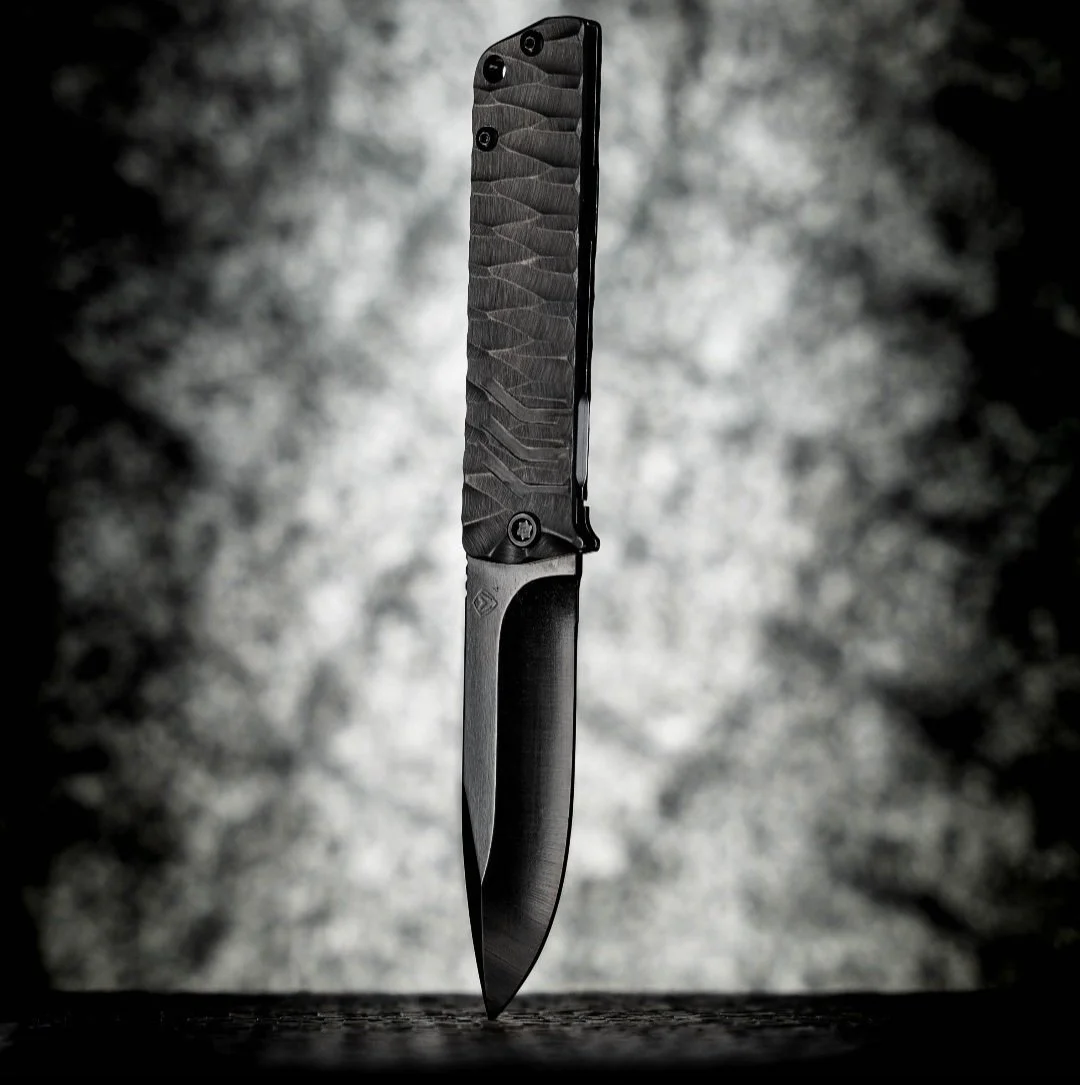 A black folding knife with a textured handle, standing upright on a dark surface against a blurry, light gray background.
