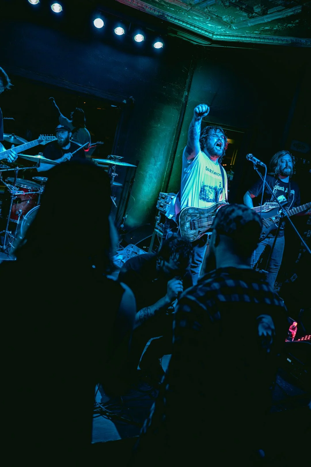 A live band performing on stage in a dimly lit venue, with musicians playing guitars and drums, and audience members watching, illuminated by colorful stage lighting.
