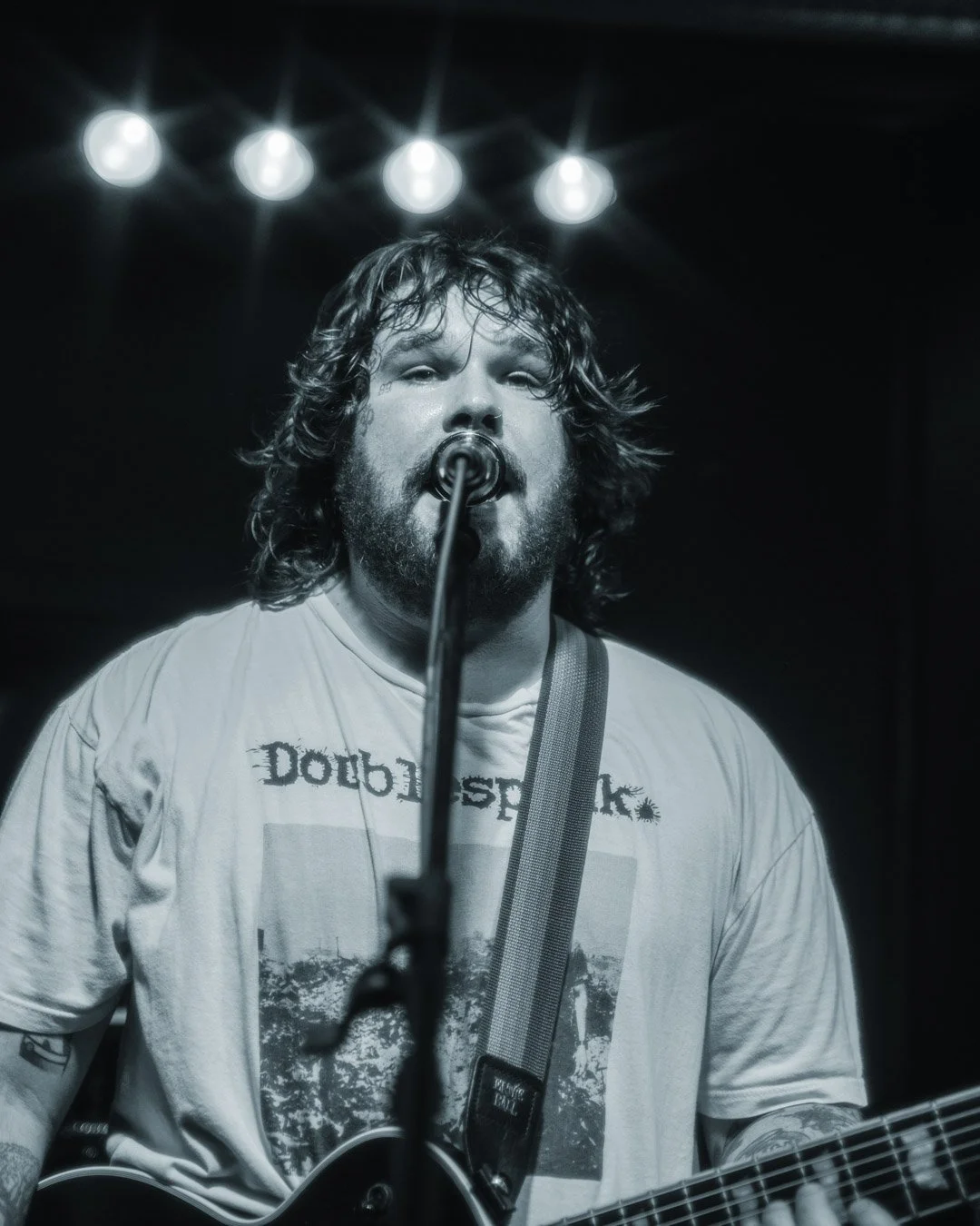 A bearded man with wavy hair performing on stage, singing into a microphone while playing an acoustic guitar, with four bright stage lights overhead.
