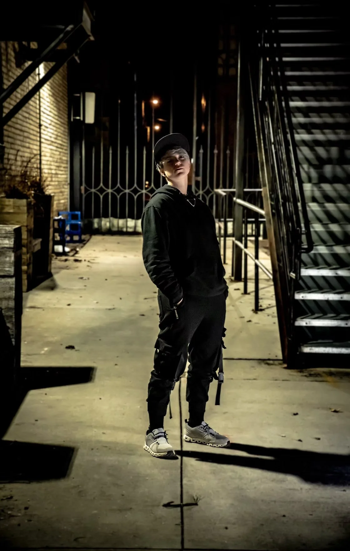 A young man stands in a dimly lit alley at night, wearing a black hoodie, black cargo pants, white sneakers, and a black cap, with his hands in his pockets and a leash hanging from his waist.