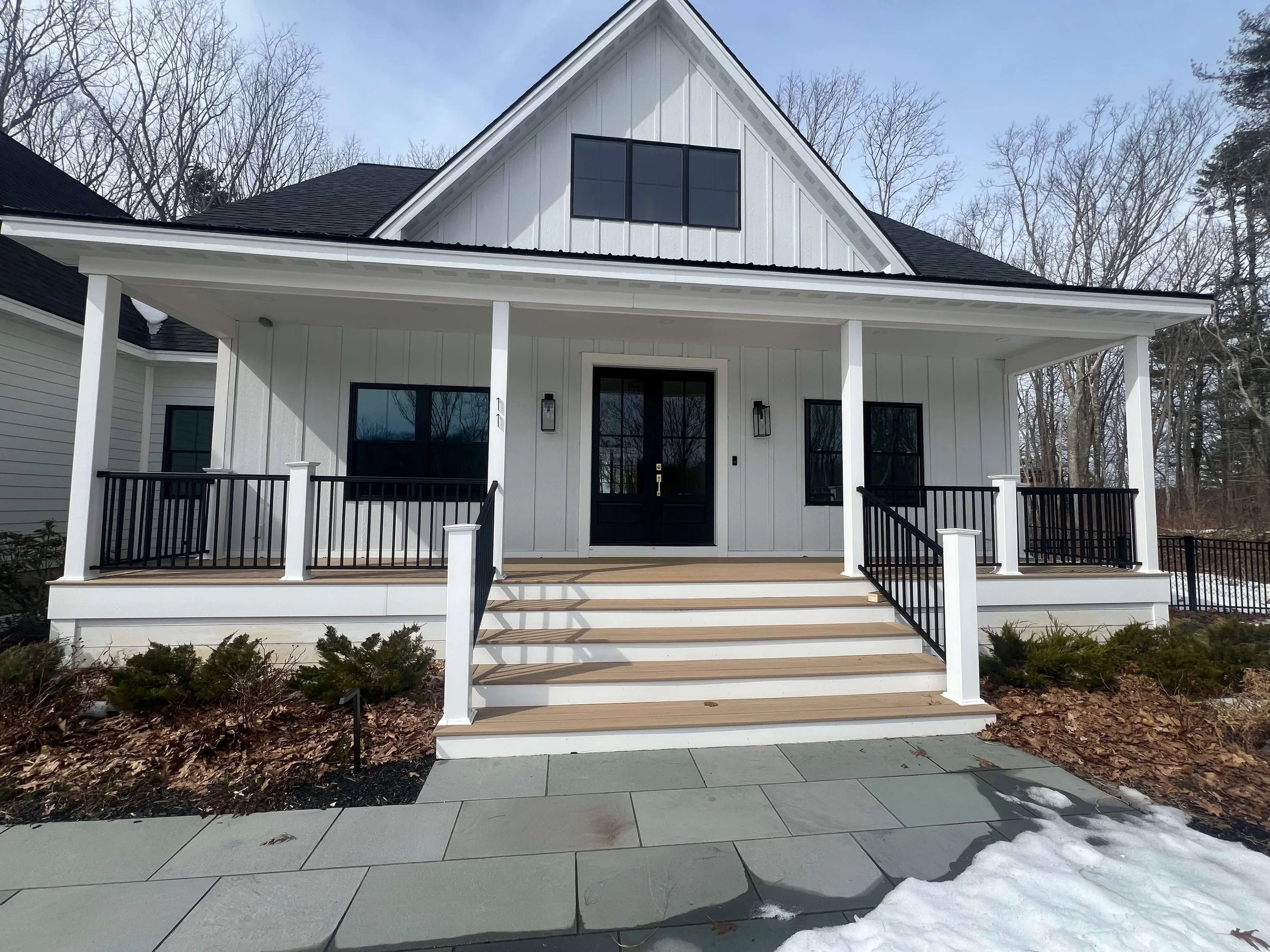 Front view of a modern white house with a front porch, black railing, black front door, large upper window, and snow on the ground.