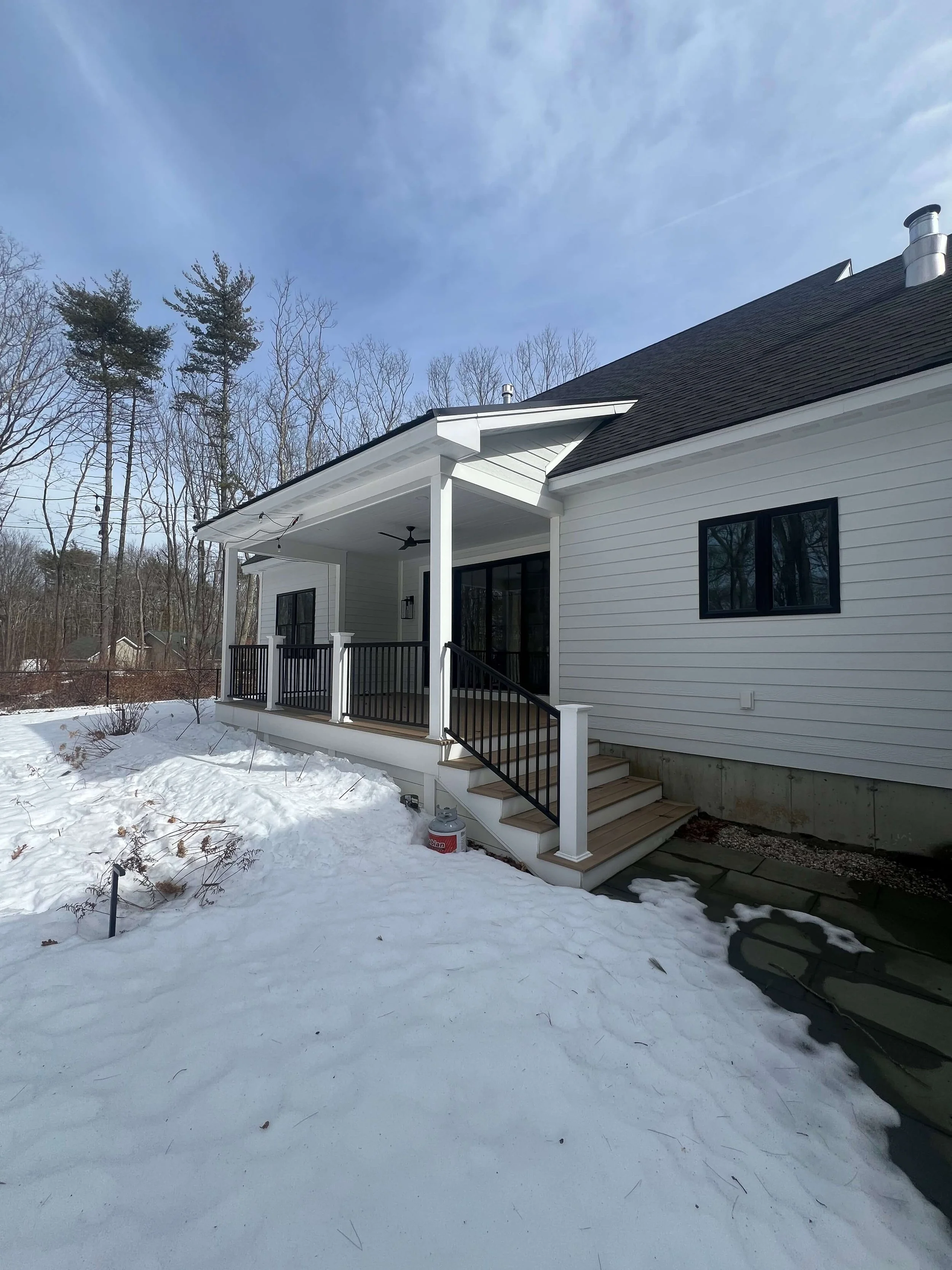 Backyard view of a white house with a small porch, black railing, steps leading up to the porch, and snow-covered ground.