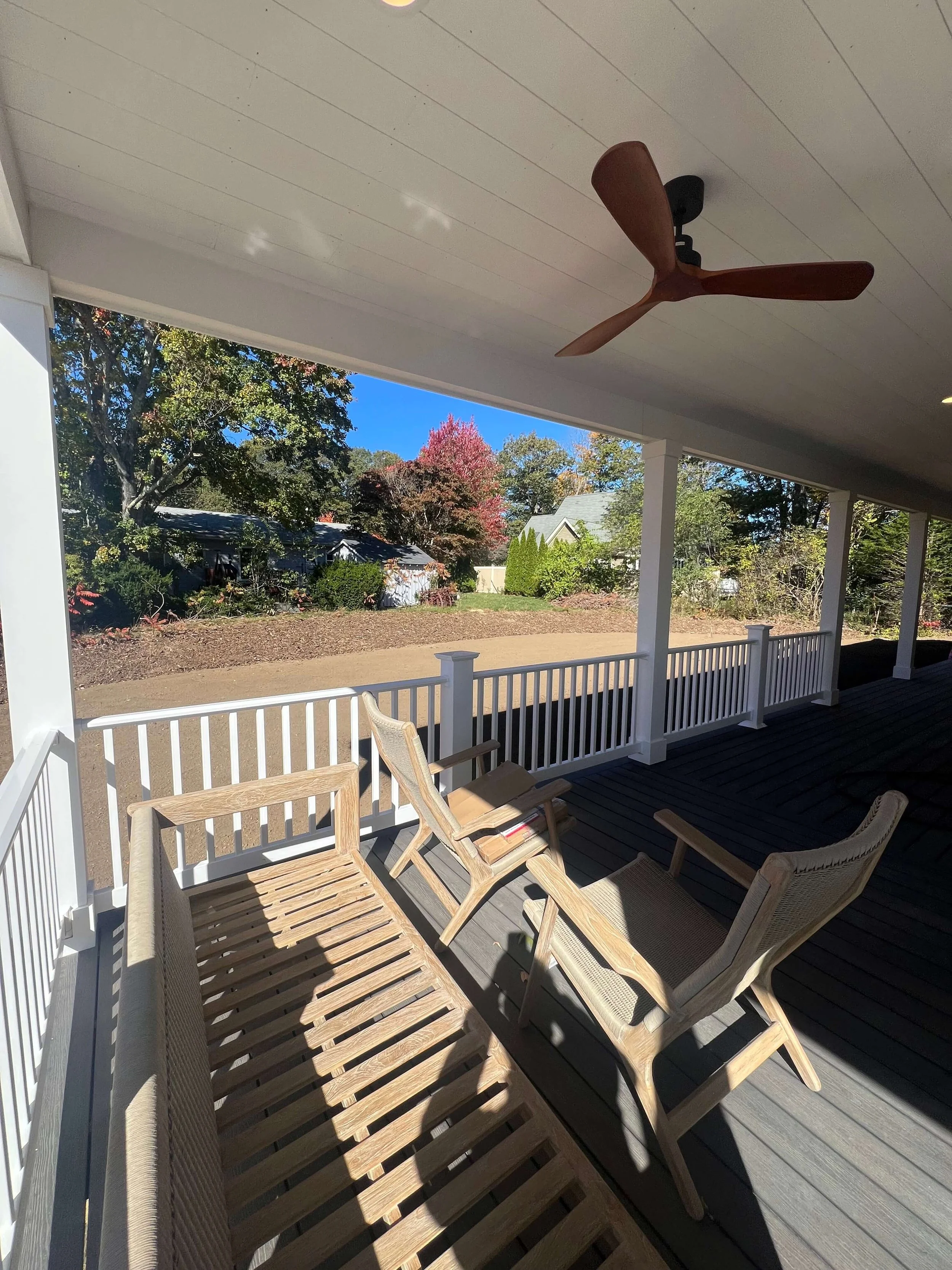 A covered porch with wooden furniture, including chairs and a bench, overlooking a garden with trees and a house in the background. There is a ceiling fan overhead.