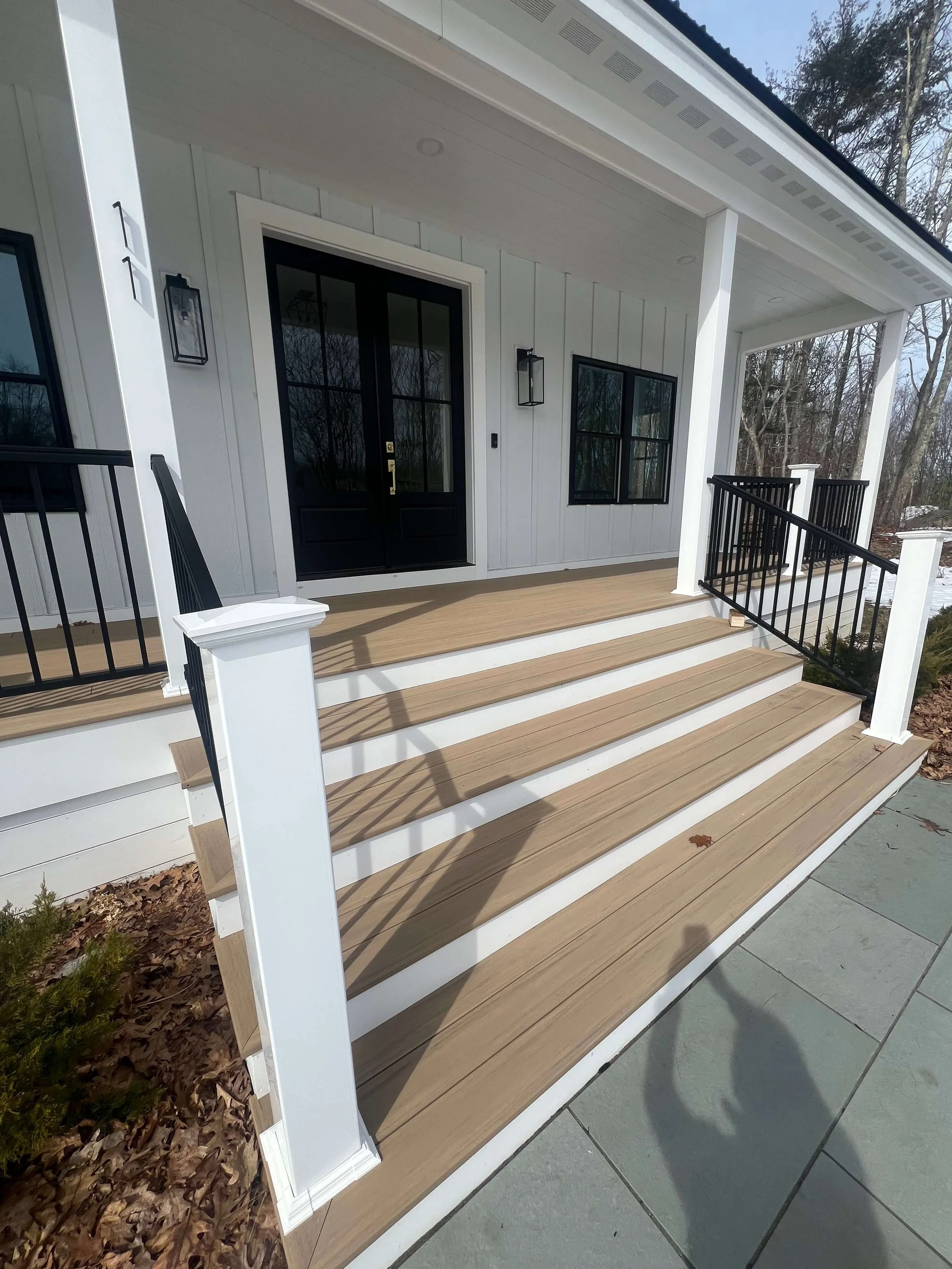 Front porch of a modern house with wooden steps, white railing posts, black railings, black door with glass panels, and two black window frames, with leafless trees in the background.