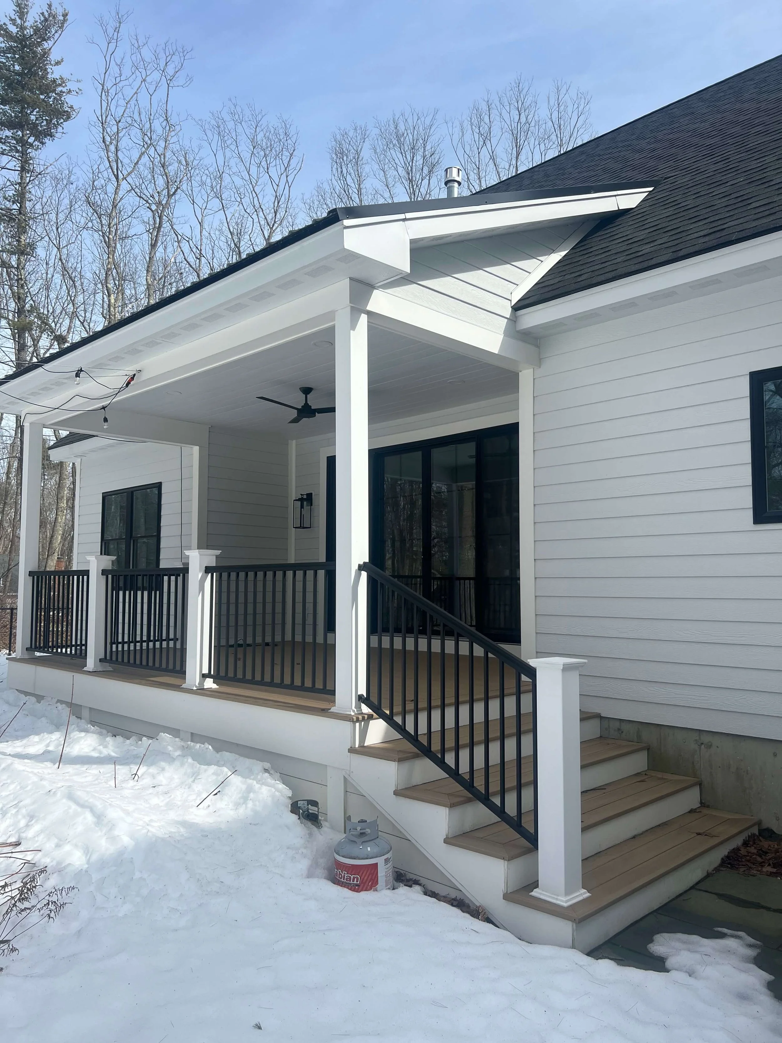 Back porch of a white house with black railings, stairs leading down to snow-covered ground, and a propane tank nearby. The porch has a ceiling fan and a light fixture, with string lights hanging from the roof.