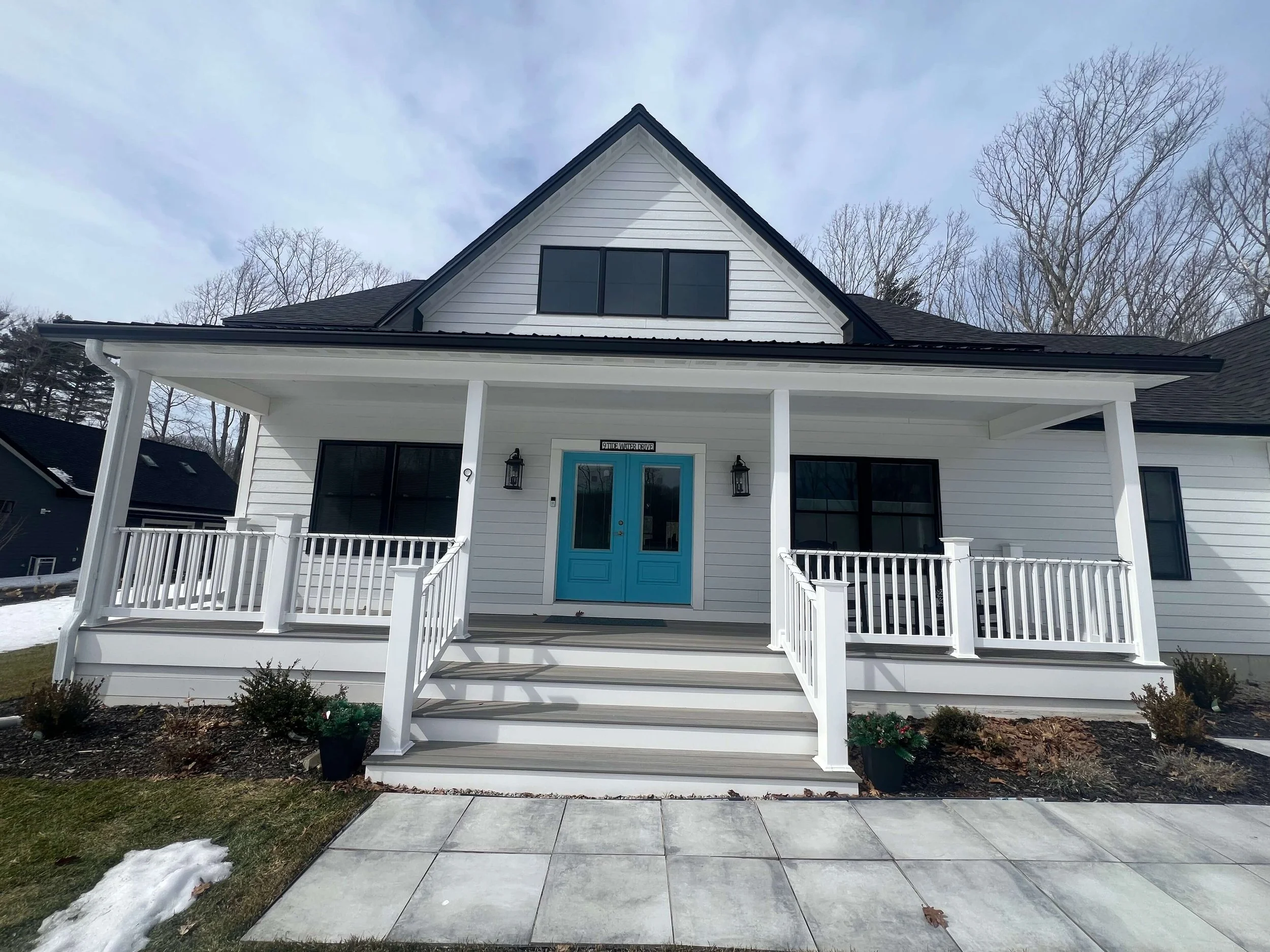 Front view of a modern white house with a front porch, blue double doors, and black window frames, surrounded by a small garden and some snow on the ground.