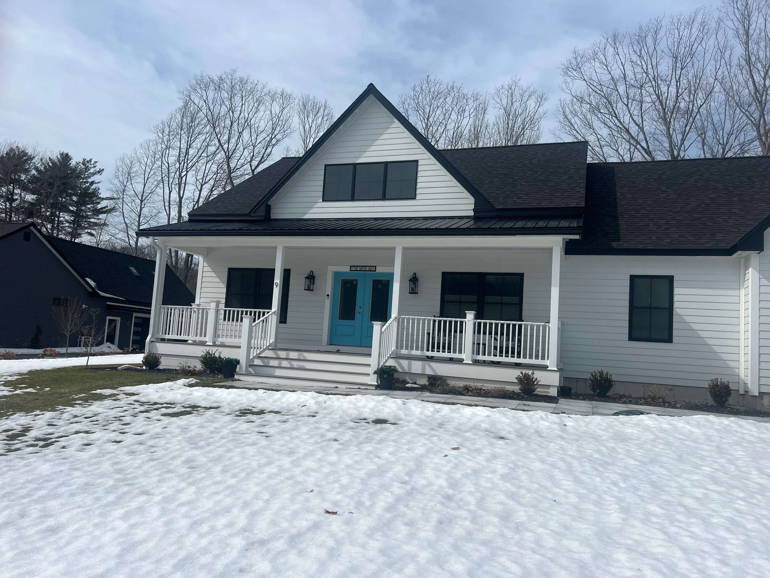 Modern white house with black roof and blue front door, snow-covered lawn, trees in background.