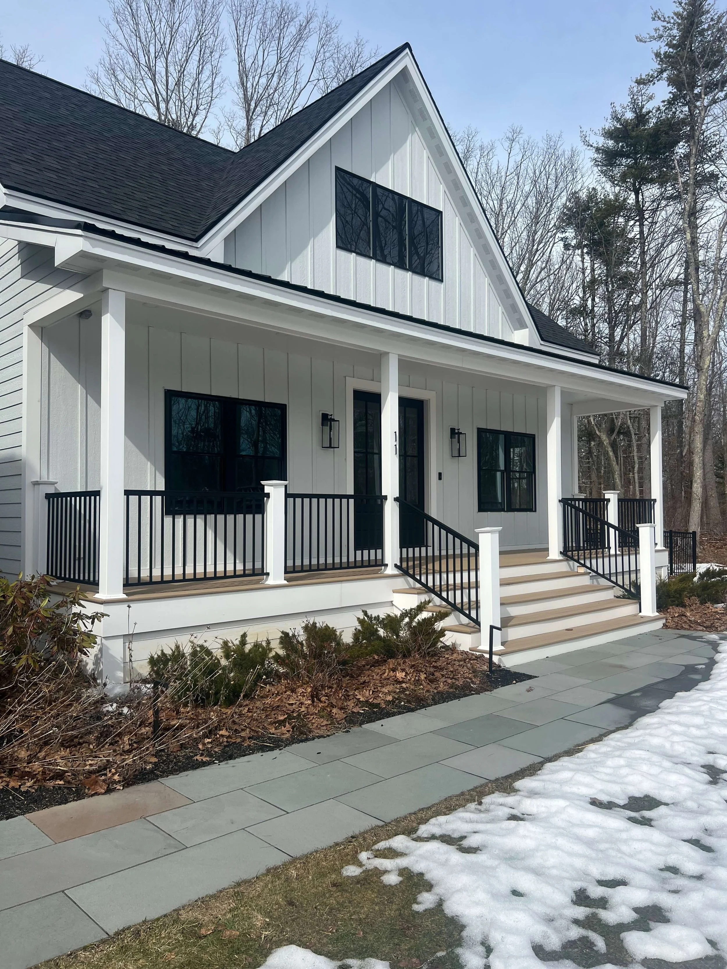 Front view of a modern white house with black accents, a porch with black railing and steps, and a snow-covered lawn.