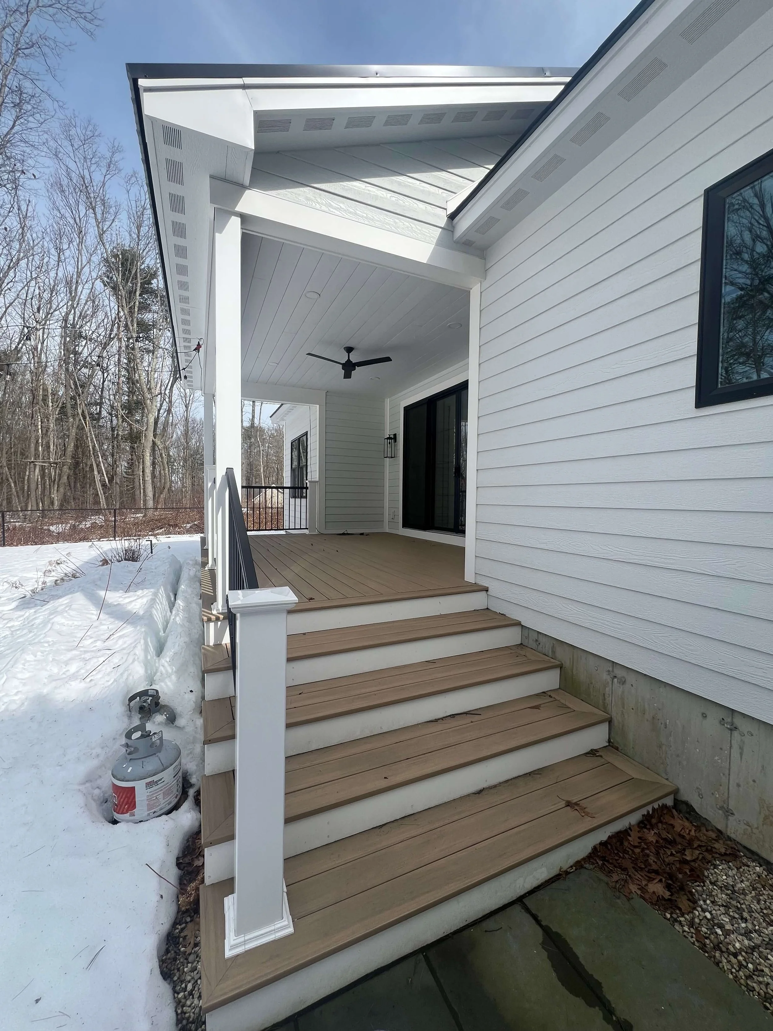 Newly constructed white house with wooden deck and stairs, black sliding glass doors, a ceiling fan, and snow on the ground outside.