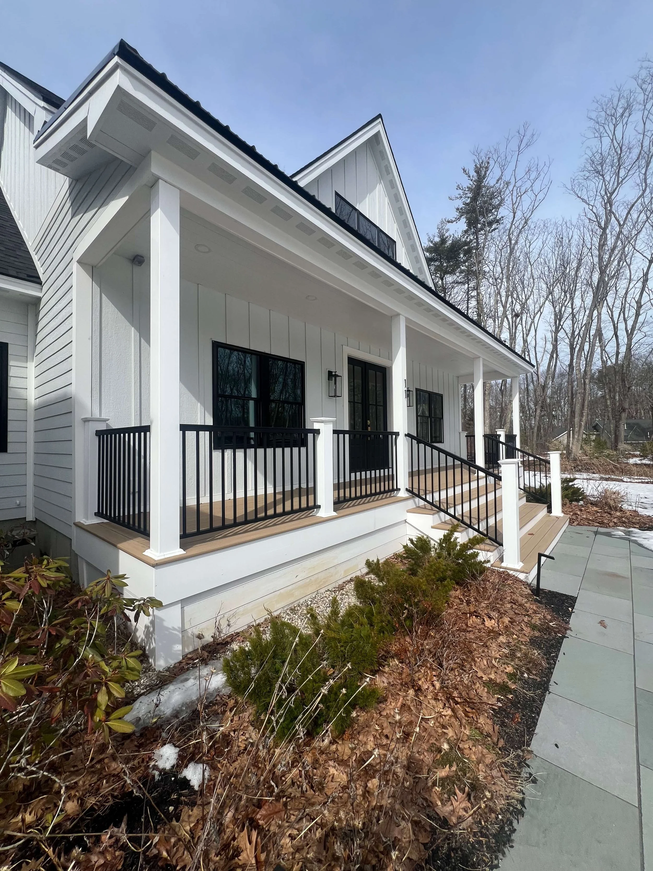 Front porch of a modern white house with black railings, steps leading up to a black door, and surrounding leafless trees in winter.