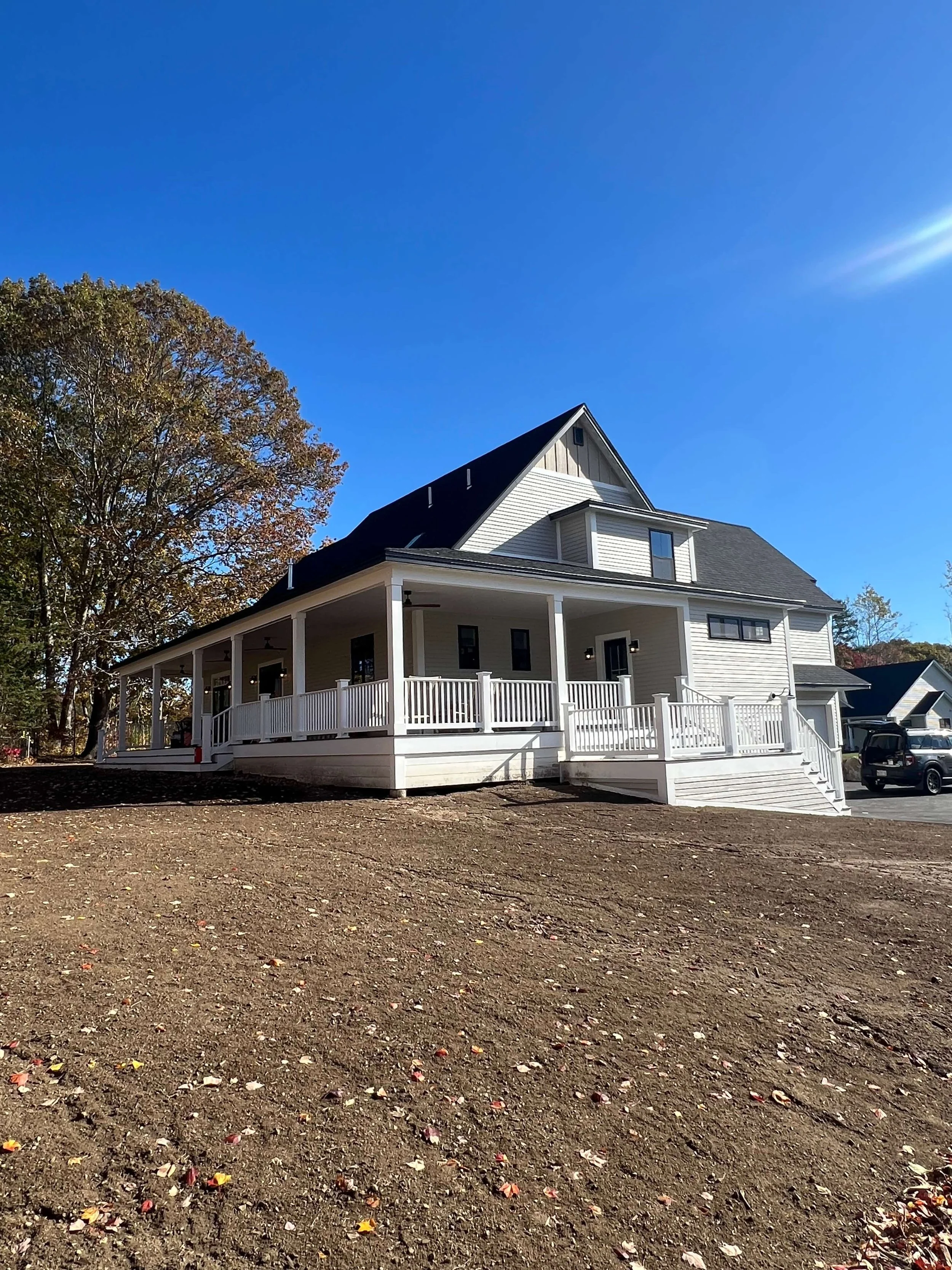 A large white house with a wraparound porch and black roof, set against a bright blue sky with a few clouds. There is a large tree with autumn leaves on the left and a parking lot with a few vehicles on the right.