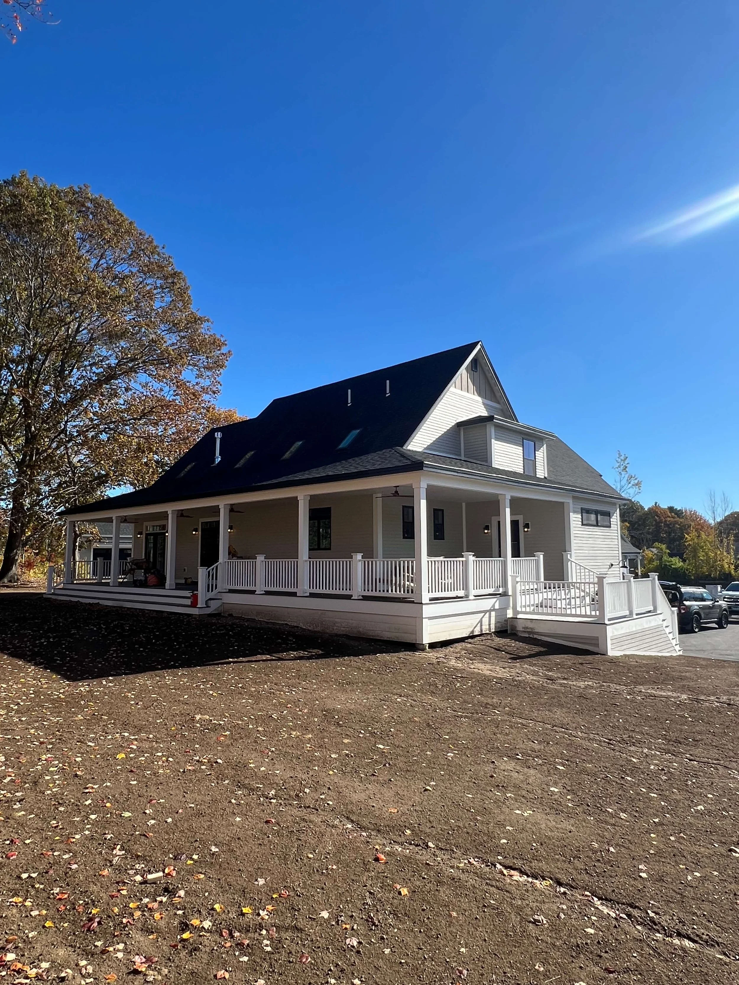 Newly constructed two-story house with a large wrap-around porch, black roof, white siding, and a small set of stairs leading to the porch, under a clear blue sky.