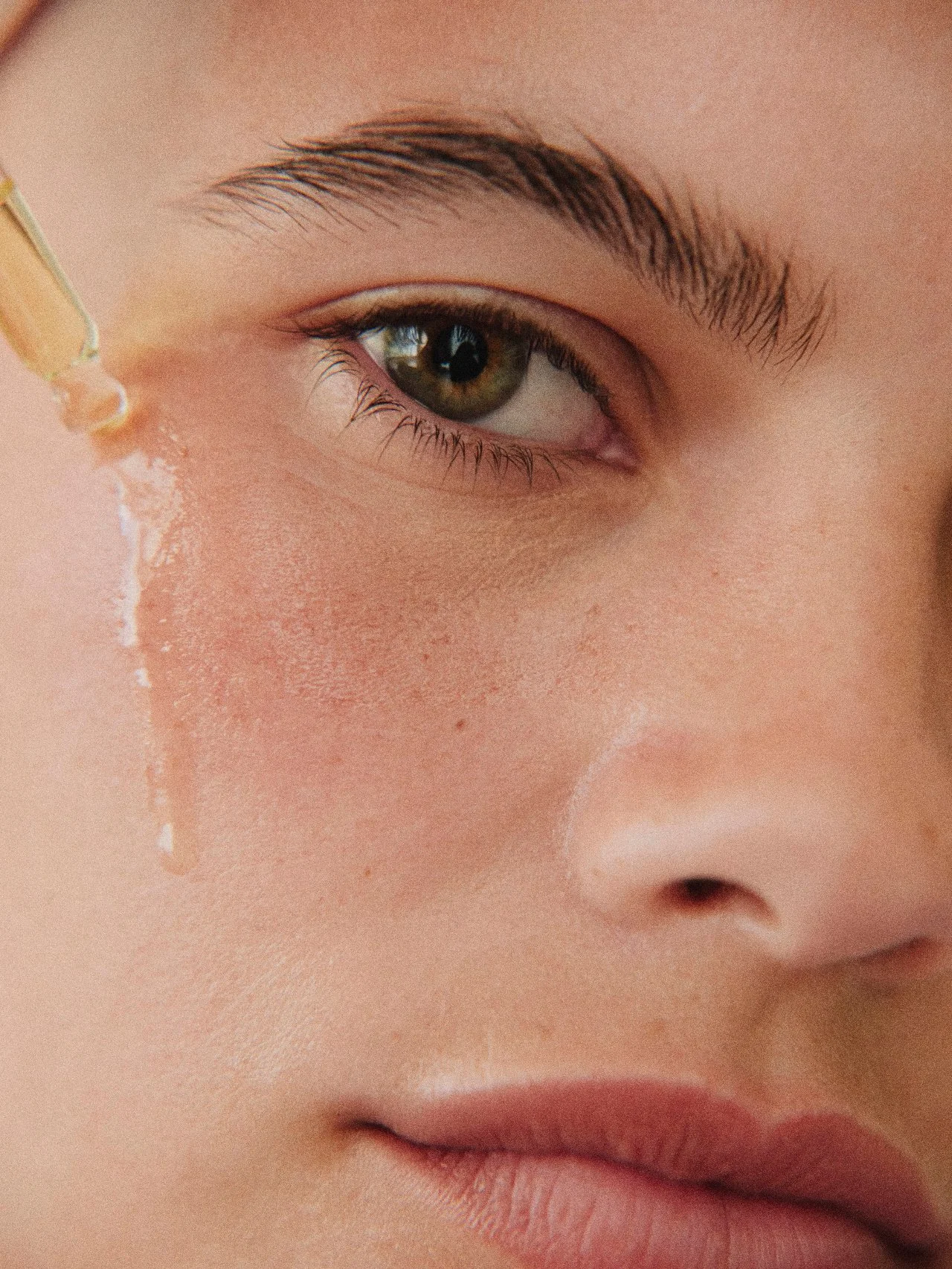Close-up of a person's eye with a dropper releasing a liquid near the eyelid, showing detailed skin texture and facial features.