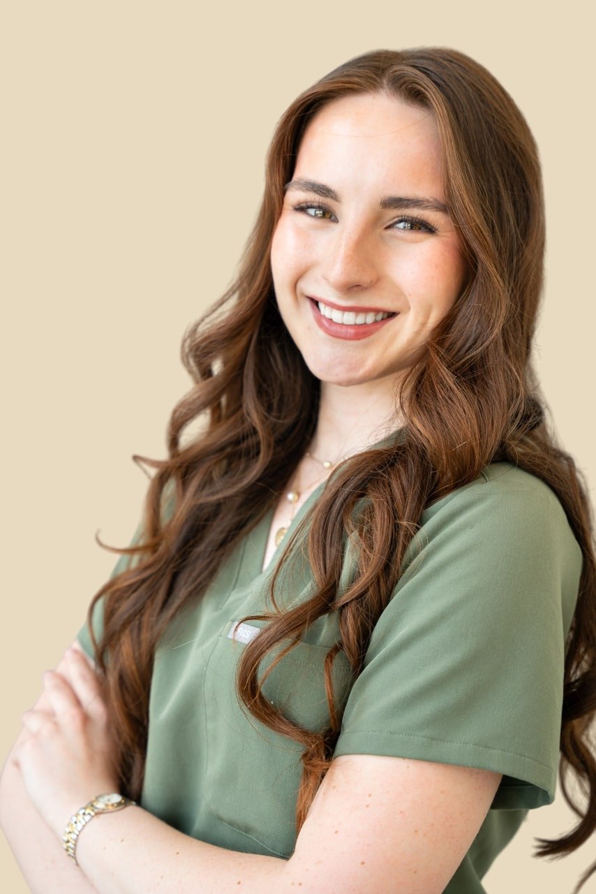 A young woman with long, wavy brown hair smiling and wearing a green scrub top, with crossed arms, standing against a beige background.
