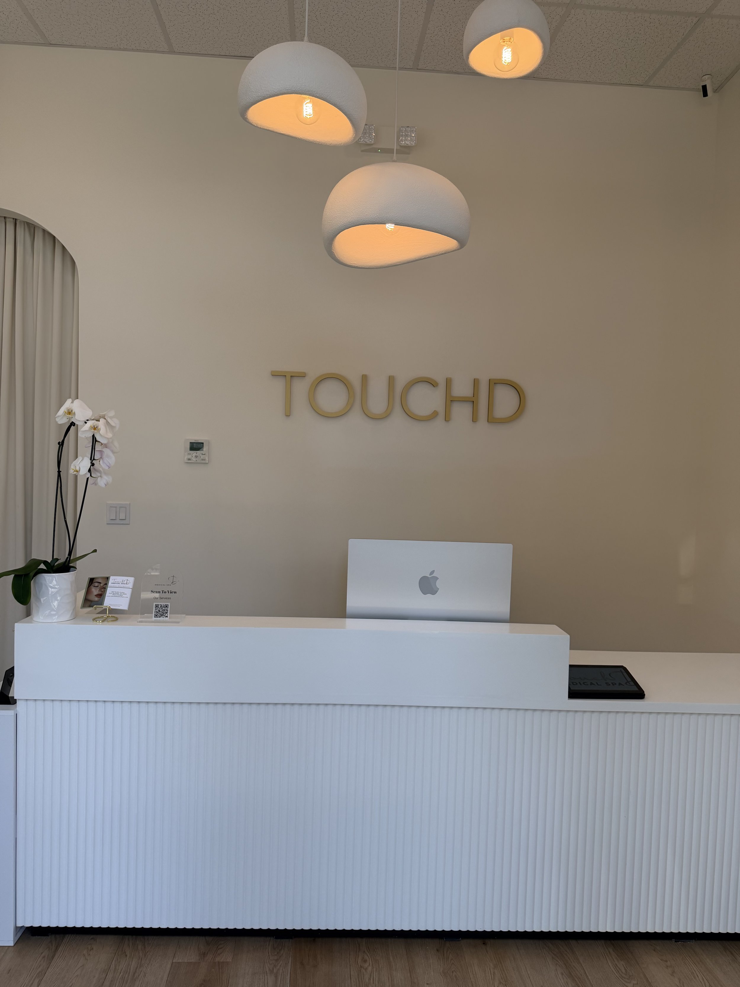 An interior reception desk area with a white counter, a tablet device, a potted orchid plant, and a wall-mounted display sign. Behind the desk there are three modern pendant lights hanging from the ceiling and a wall with the text 'TOUCHD' displayed in gold lettering.