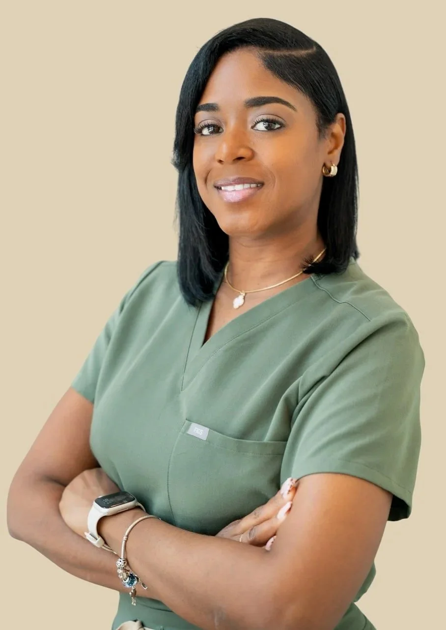 A woman wearing green medical scrubs with arms crossed, smiling, and accessorized with jewelry, against a beige background.