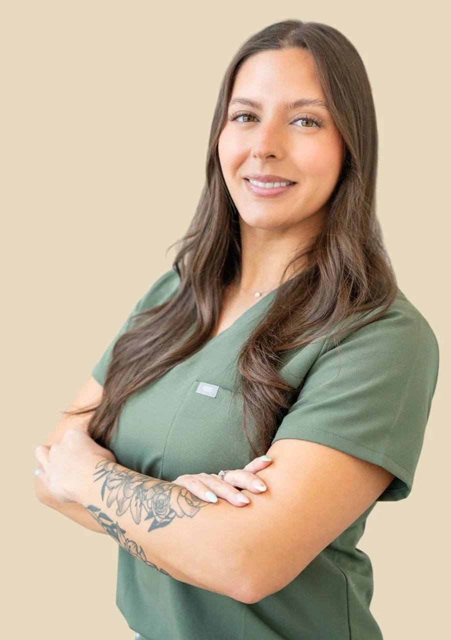 A woman wearing green medical scrubs with a name tag. She has long brown hair, a tattoo on her right forearm, and is smiling with arms crossed against a beige background.