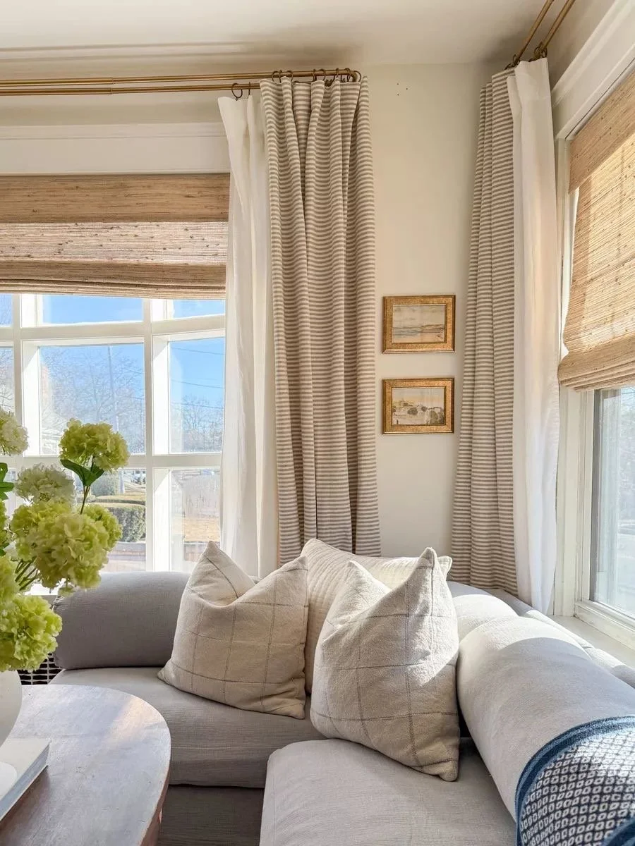 A cozy sitting area with beige and white striped curtains, a white sofa with beige pillows, and a window with natural sunlight.