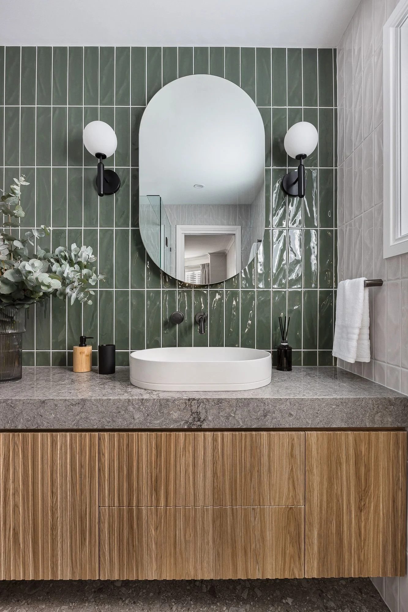 Modern bathroom vanity with a stone countertop, white vessel sink, and a large oval mirror. Green tiled wall behind the vanity features two black wall sconces with white glass shades. A small plant, soap dispenser, and reed diffuser decorate the countertop. A towel hangs on a bar to the right, and part of a window is visible.