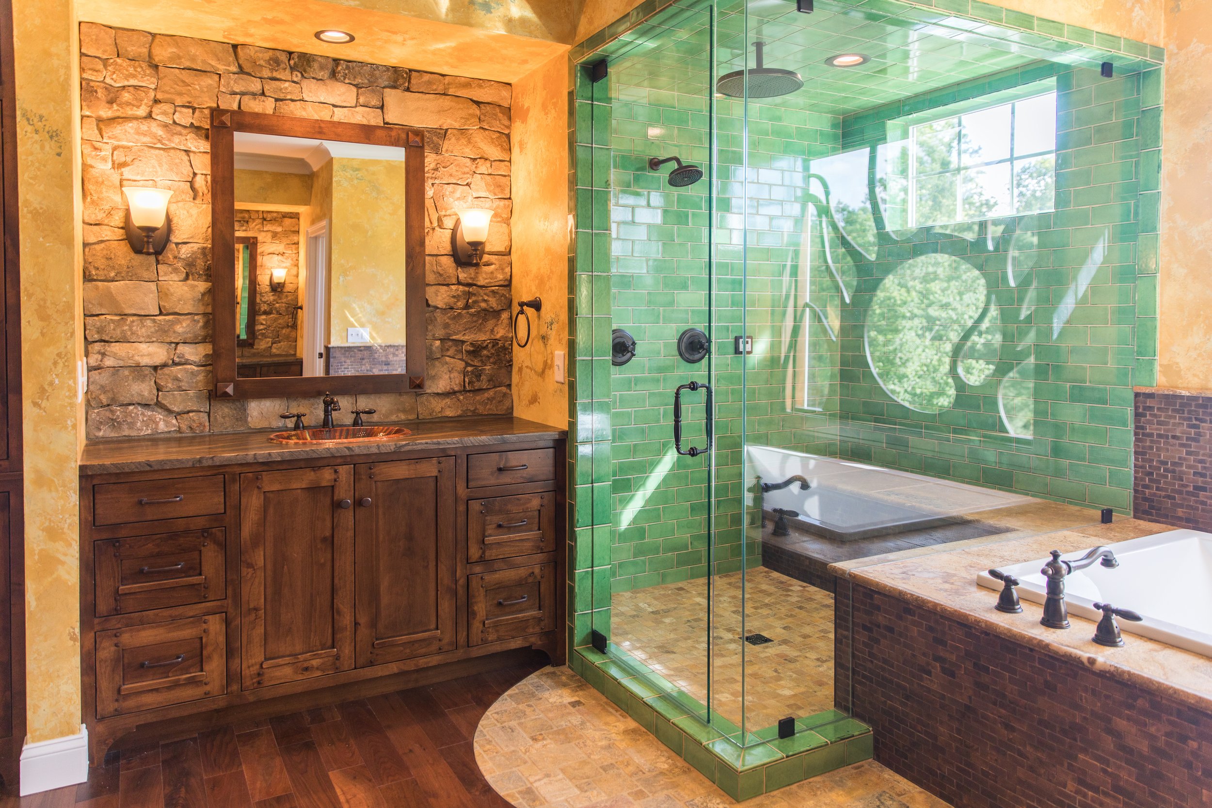 Bathroom with a stone wall mirror, wooden vanity, green tiled shower with glass door, and bathtub with brown tiled exterior.