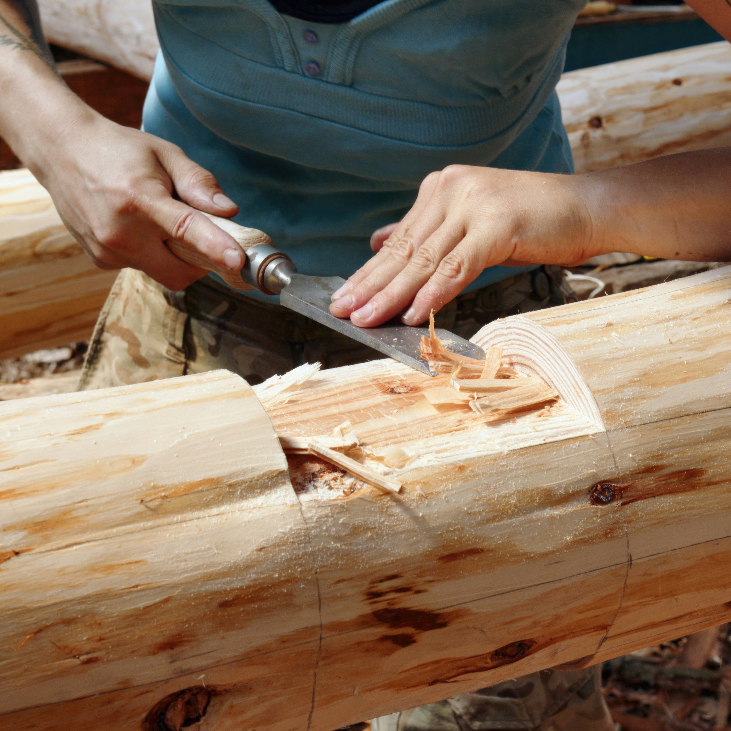 Person shaping or carving a piece of wood with a hand-held rotary tool or grinder, working on a wooden structure or furniture piece.