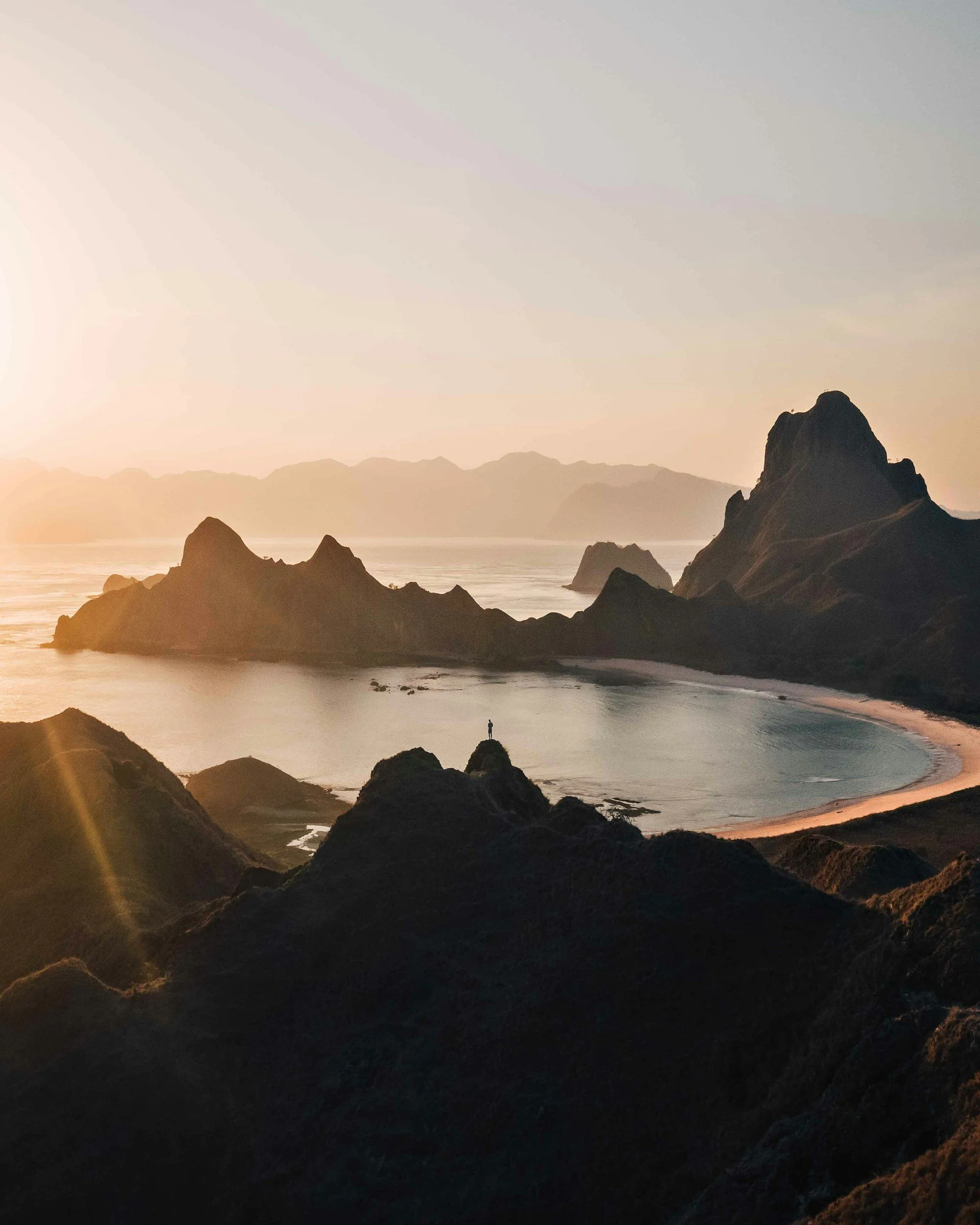 A person standing on a rock overlooking a bay with mountains in the background and the setting sun.