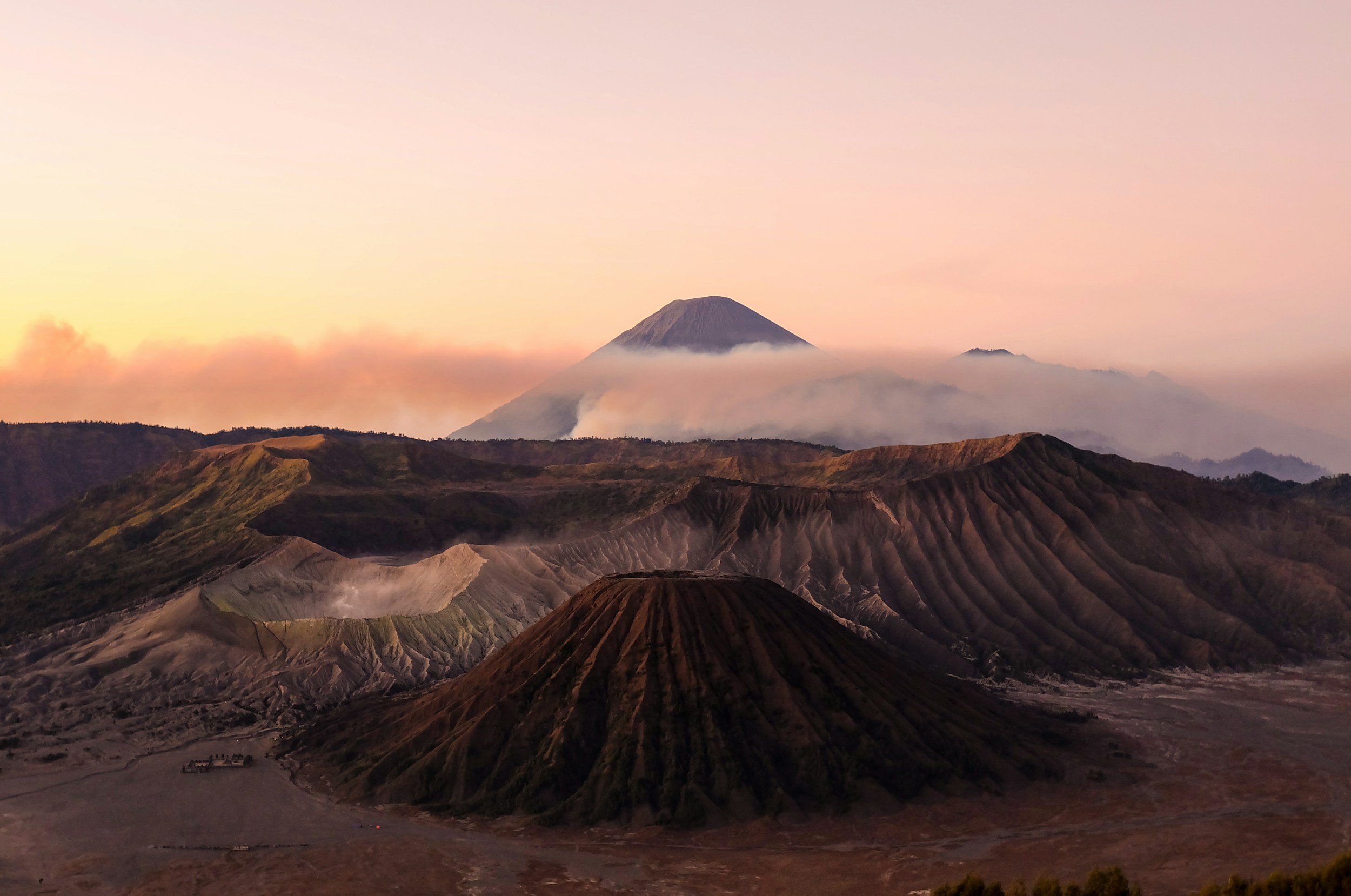 A view of volcanic craters and Mount Semeru in the background at sunrise, with volcanic ash and smoke visible.