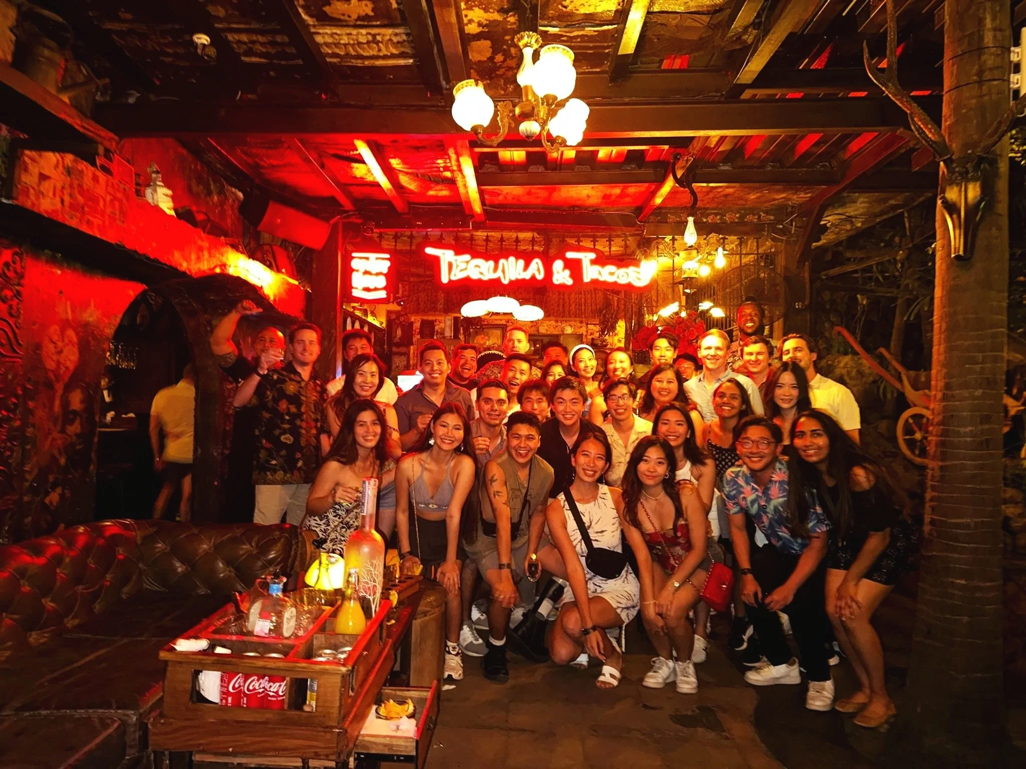 A large group of friends posing together for a photo inside a Mexican-themed restaurant or bar with a neon sign that says 'Tequila & Tacos' behind them.