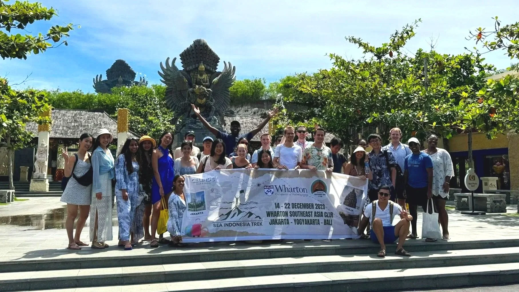 Group of people holding a banner in front of a statue with a dragon, surrounded by trees, with clear blue sky above.