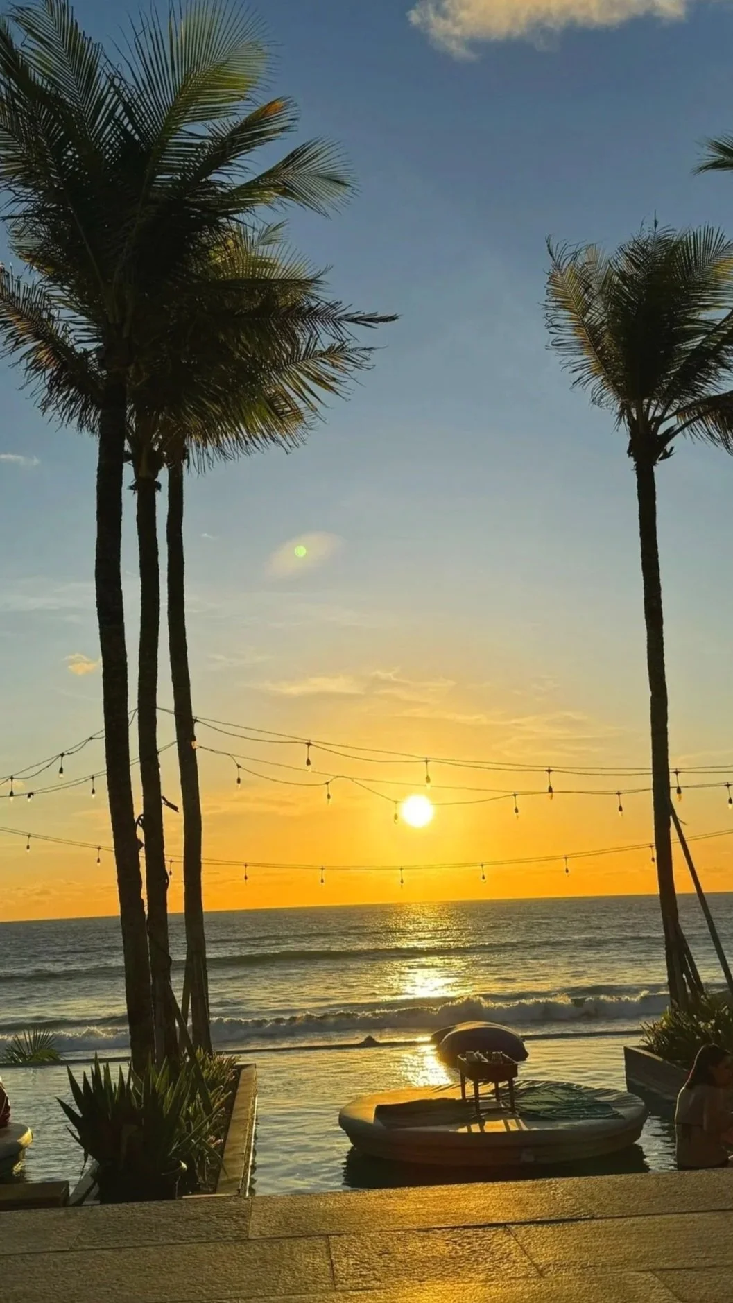 Sunset over the ocean with palm trees and string lights hanging across the sky.