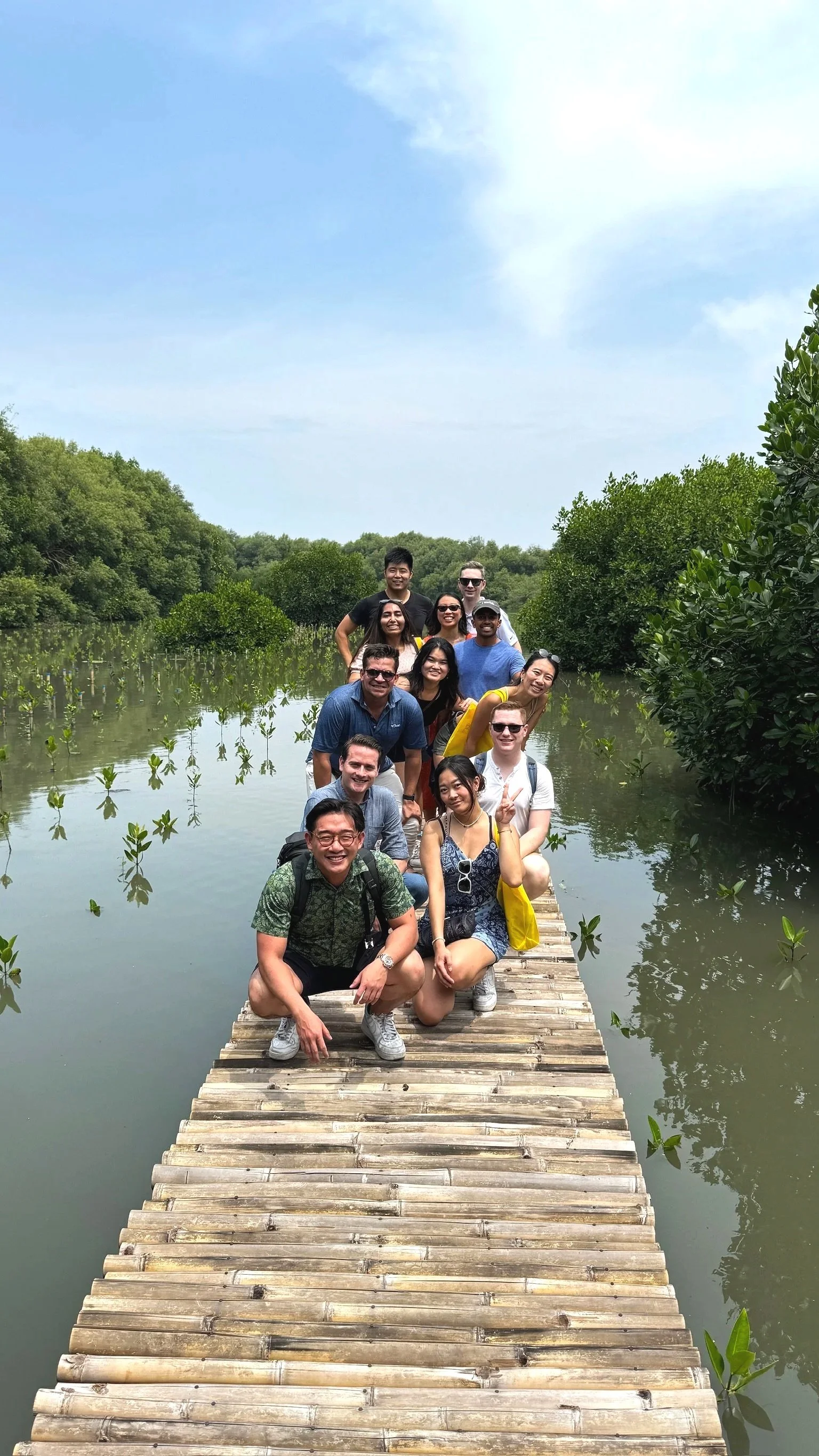 Group of people posing on a wooden dock surrounded by water with green foliage and trees, under a partly cloudy sky.