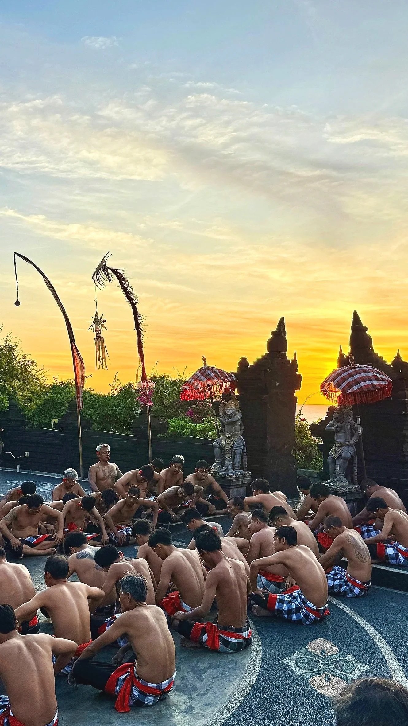 Group of men wearing traditional sarongs seated on ground during sunset at a cultural site, with statues and decorative umbrellas in the background.