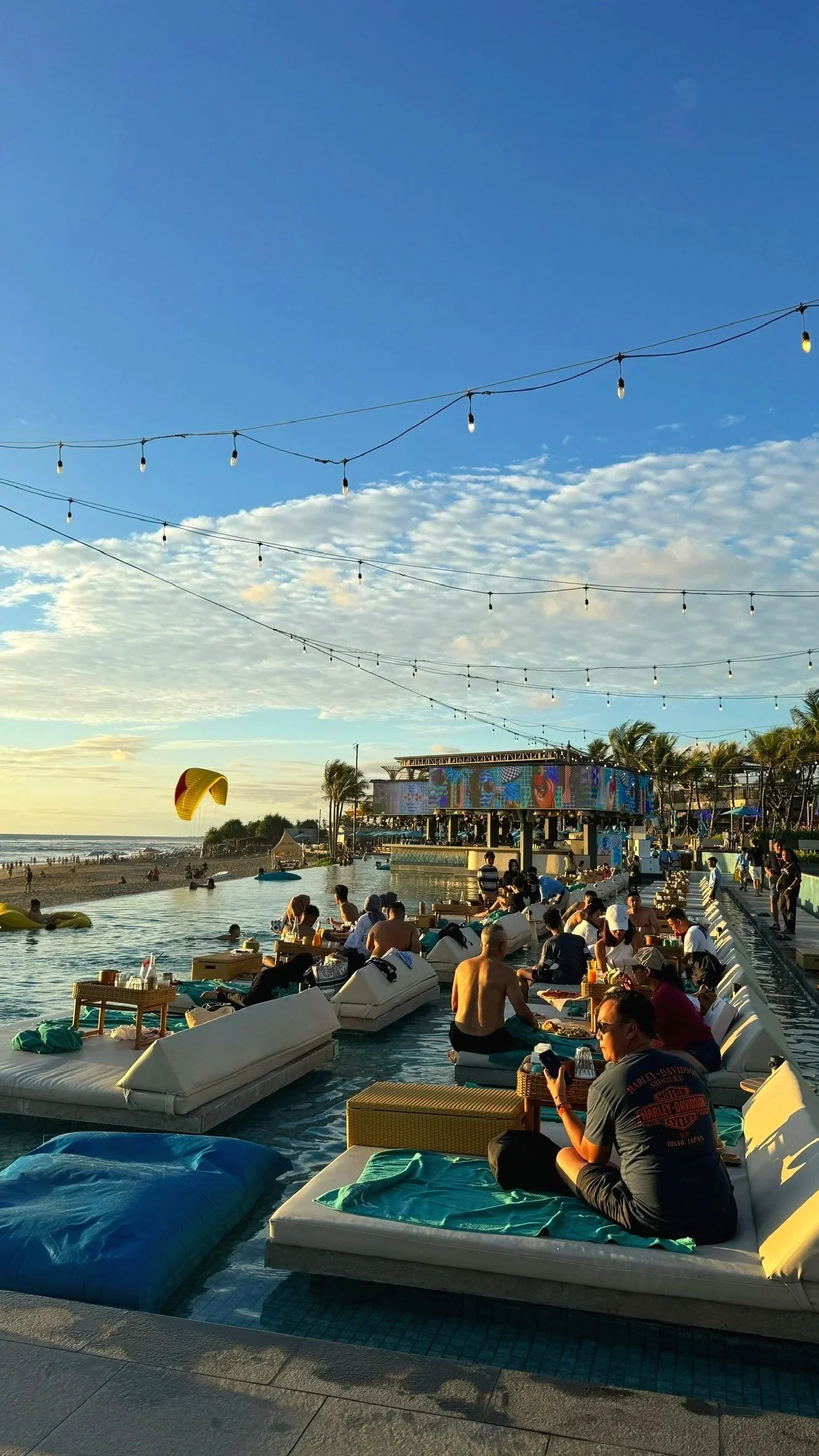 People relaxing and socializing by a poolside with cityscape and the beach in the background during sunset, with string lights overhead.