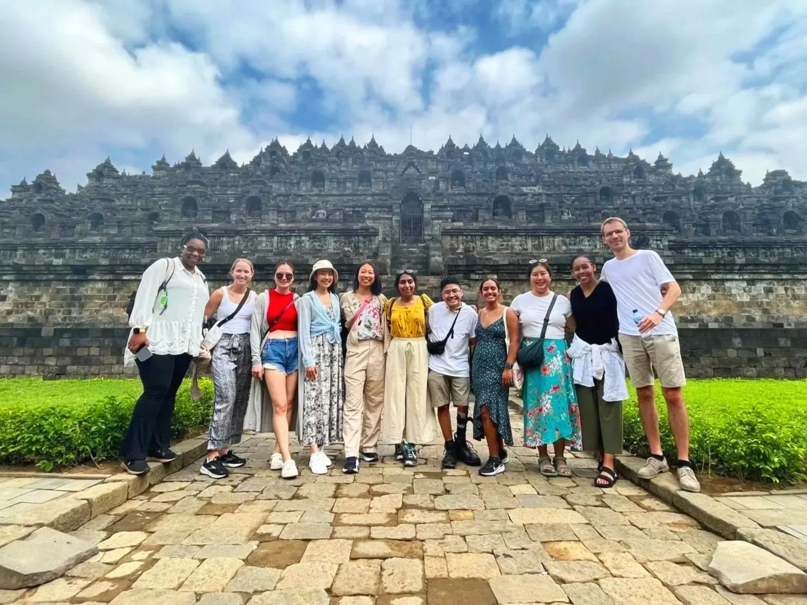 Group of twelve diverse tourists posing in front of a large ancient stone temple, with a cobblestone path and green bushes in the foreground, and cloudy sky above.