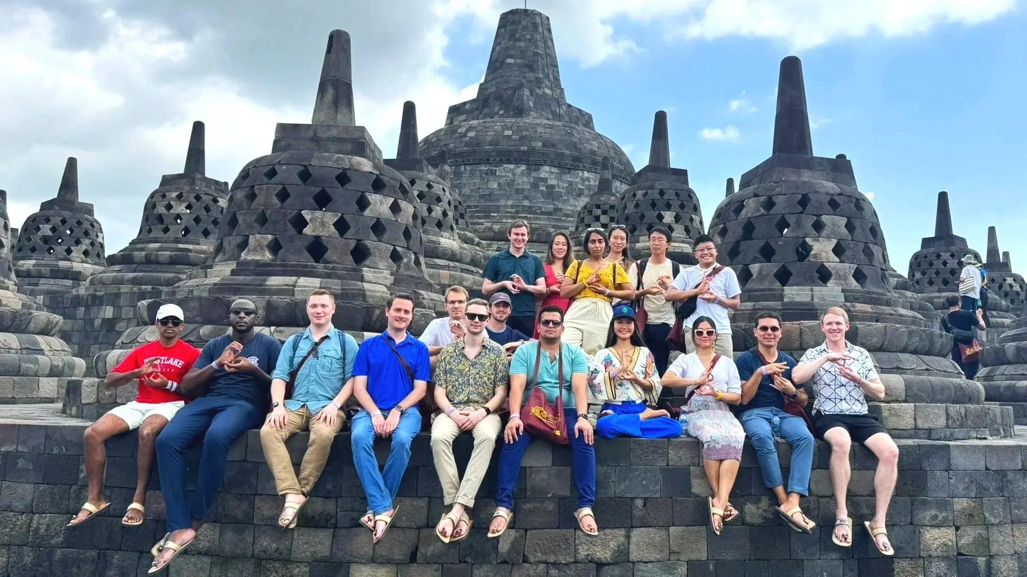 Group of tourists sitting and standing on stone steps in front of Borobudur temple, Indonesia, with a cloudy sky in the background.