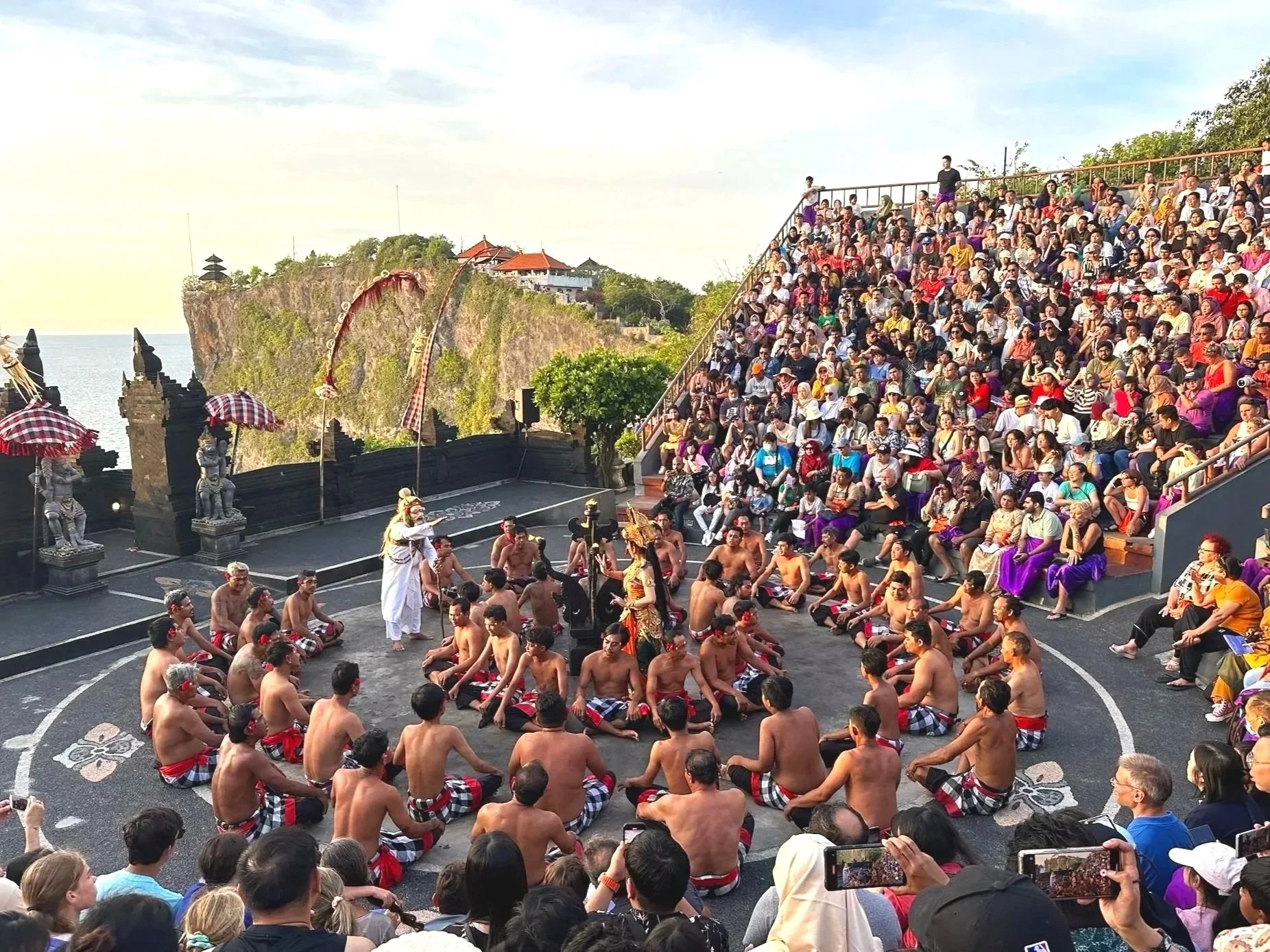 Traditional Balinese dance performance with musicians and audience by a cliffside on the beach.
