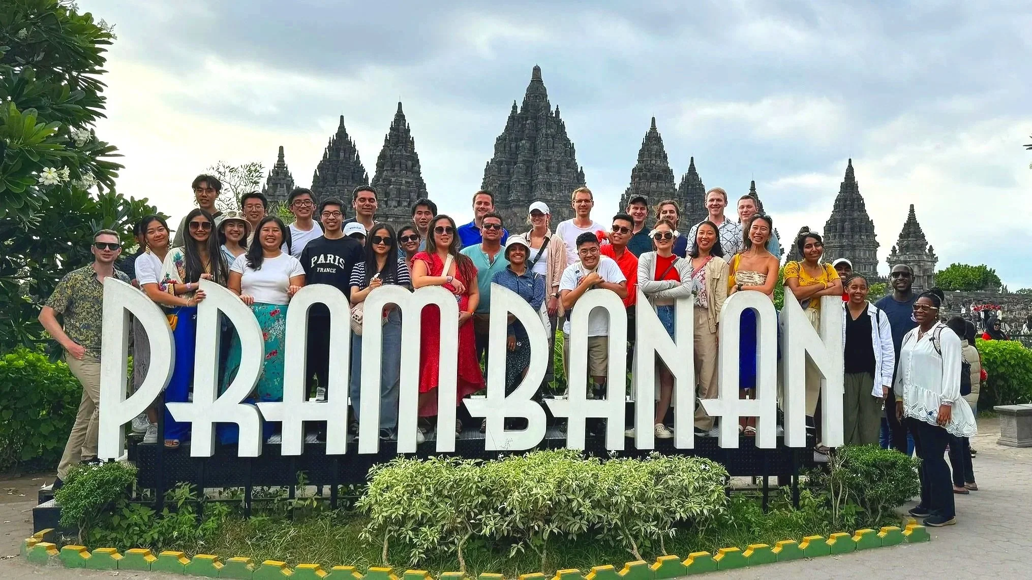 Group of tourists posing behind a large 'PRAMBANAN' sign in front of the Prambanan Temple complex in Indonesia, with ancient Hindu temples in the background.