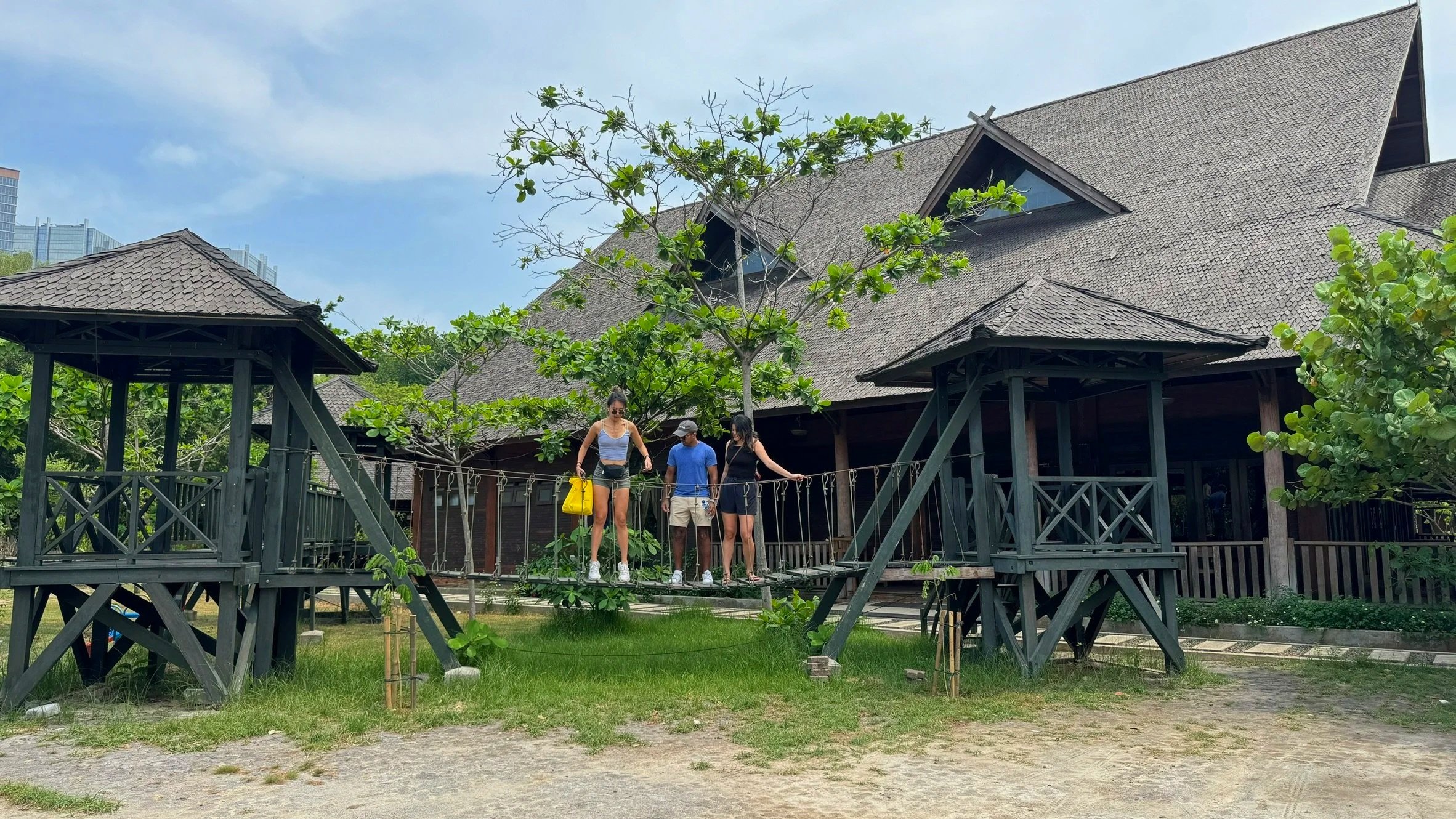 Three people standing on a suspended bridge in front of a traditional-style wooden building with a steep roof, surrounded by green trees and grass, with modern buildings visible in the background.