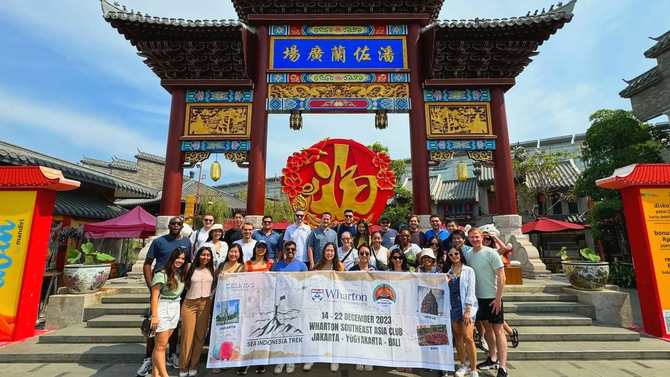 Group of tourists standing in front of a traditional Chinese gate holding a banner about an Southeast Asia trekking trip from Jakarta to Bali, with clear sky and colorful decorations.