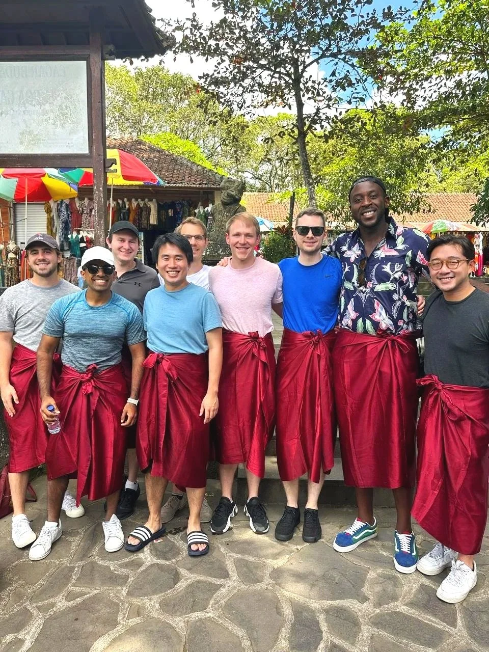 A group of eight men standing outdoors, wearing casual clothes and traditional red sarongs, smiling for a photo.