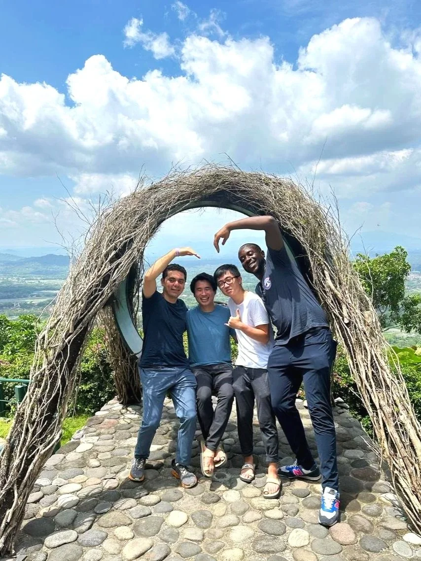 Four friends standing under a circular arch made of sticks, smiling and posing with their arms forming a heart shape, on a cobblestone platform outdoors under a blue sky with clouds.
