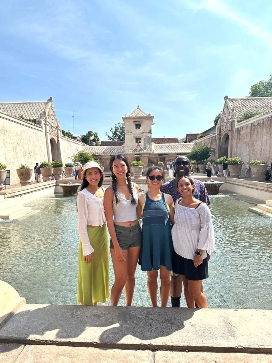 Group of five diverse women and men smiling and standing together in front of a fountain at a historical site with old walls and buildings under a clear blue sky.