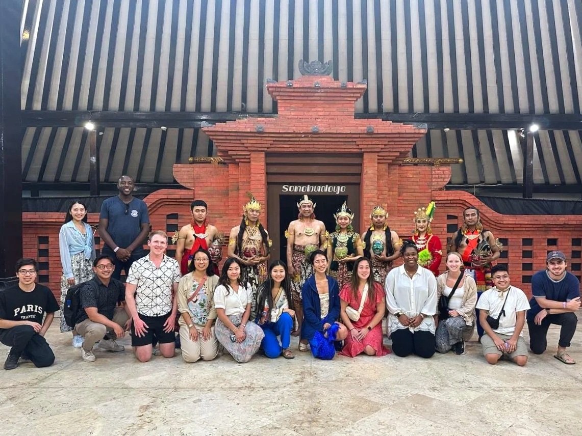 Group of diverse people posing in front of traditional Indonesian puppet theater with wooden detailed facade and intricate costumes.