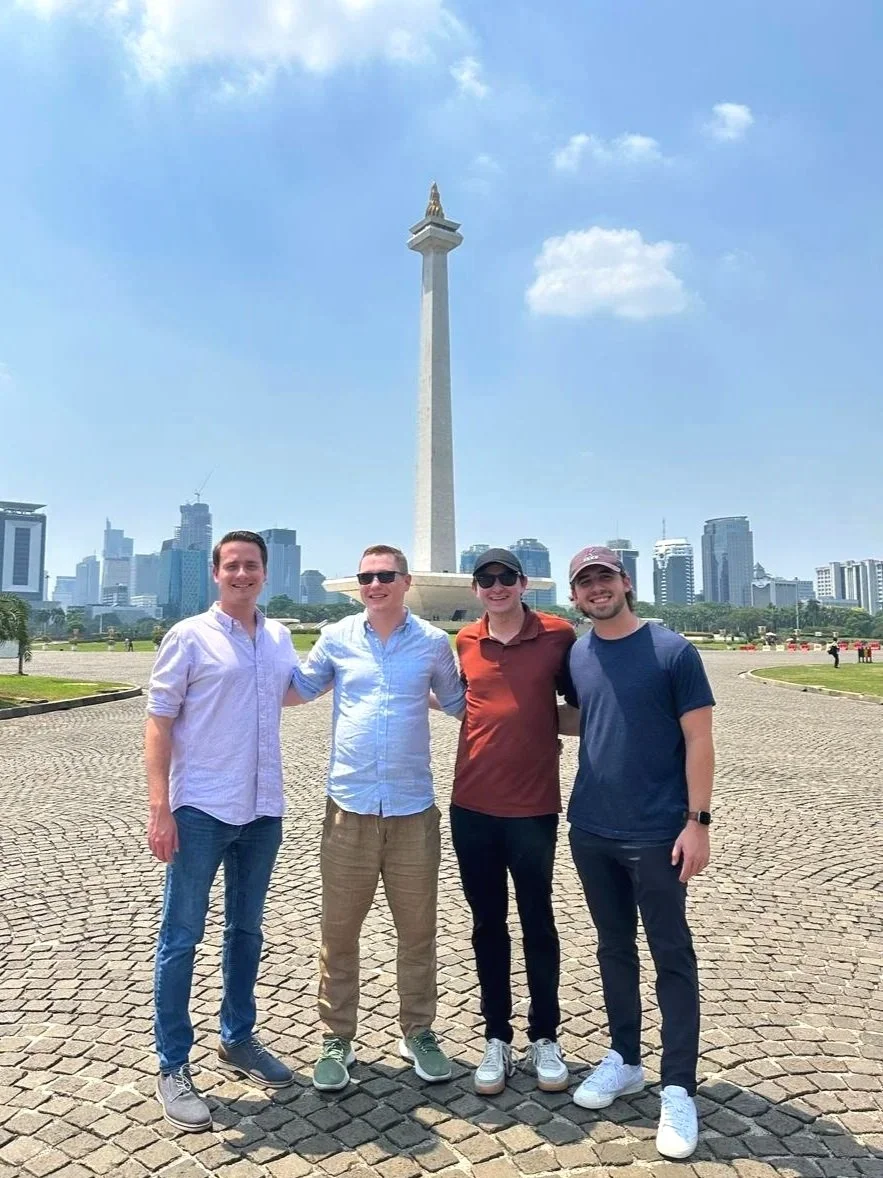 Four young men standing side by side on a paved area in front of a large monument with a city skyline in the background. The sky is mostly clear with a few clouds.