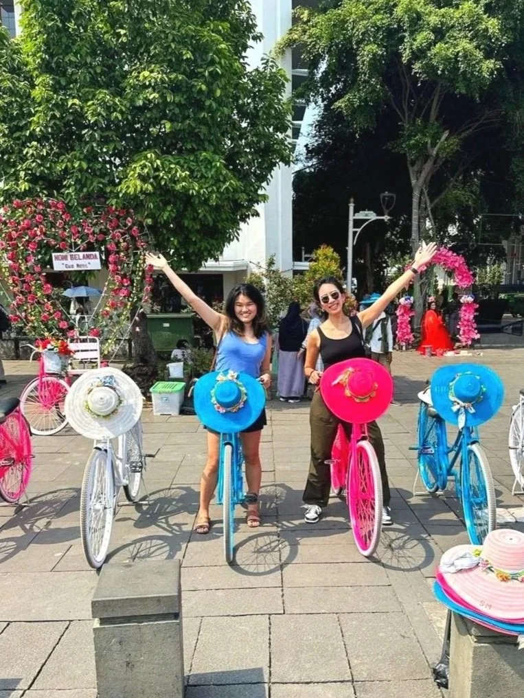 Two women happy and smiling, standing with colorful decorated bicycles in an outdoor urban square, surrounded by trees and other people, with flower wreaths and hats in the background.