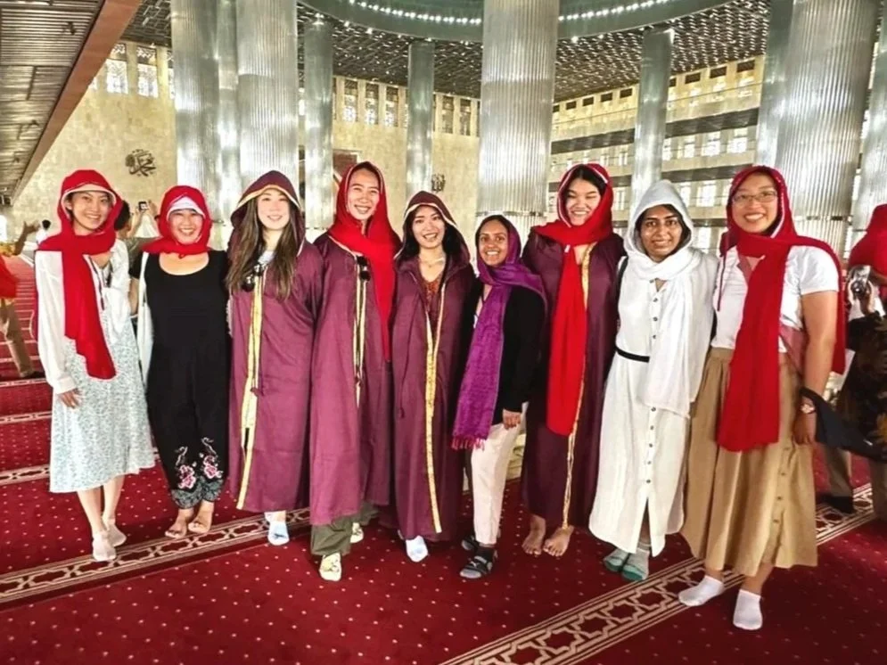 Group of nine women in colorful traditional and casual clothing, some wearing scarves and hats, standing inside a modern building with metallic columns and a red patterned carpet, posing for a photo.