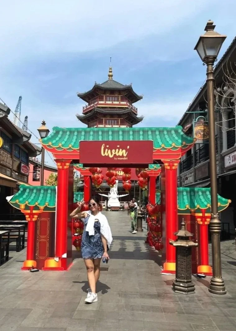 A woman walking through a Chinese-themed entrance with red pillars, green tiled roof, and red lantern decorations, leading to a pagoda-style building.